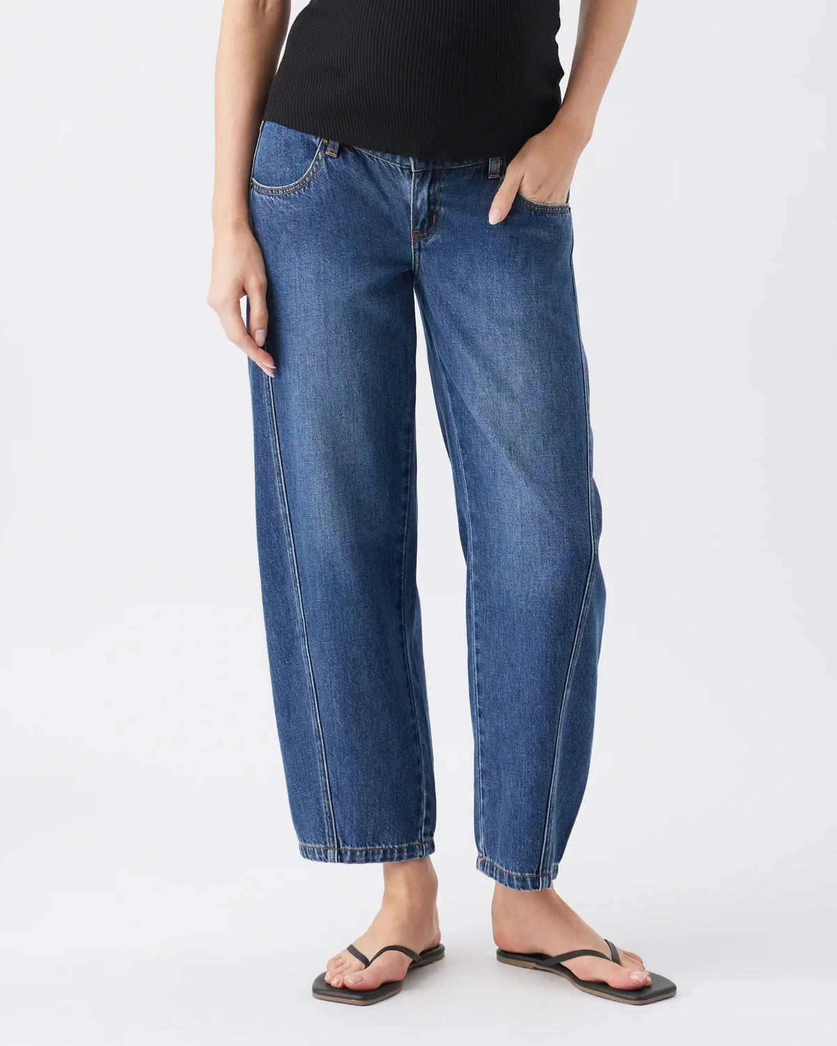 A person wearing medium-wash blue cropped jeans with a black ribbed top and black flip-flop sandals, shown from the waist down against a light gray studio background.