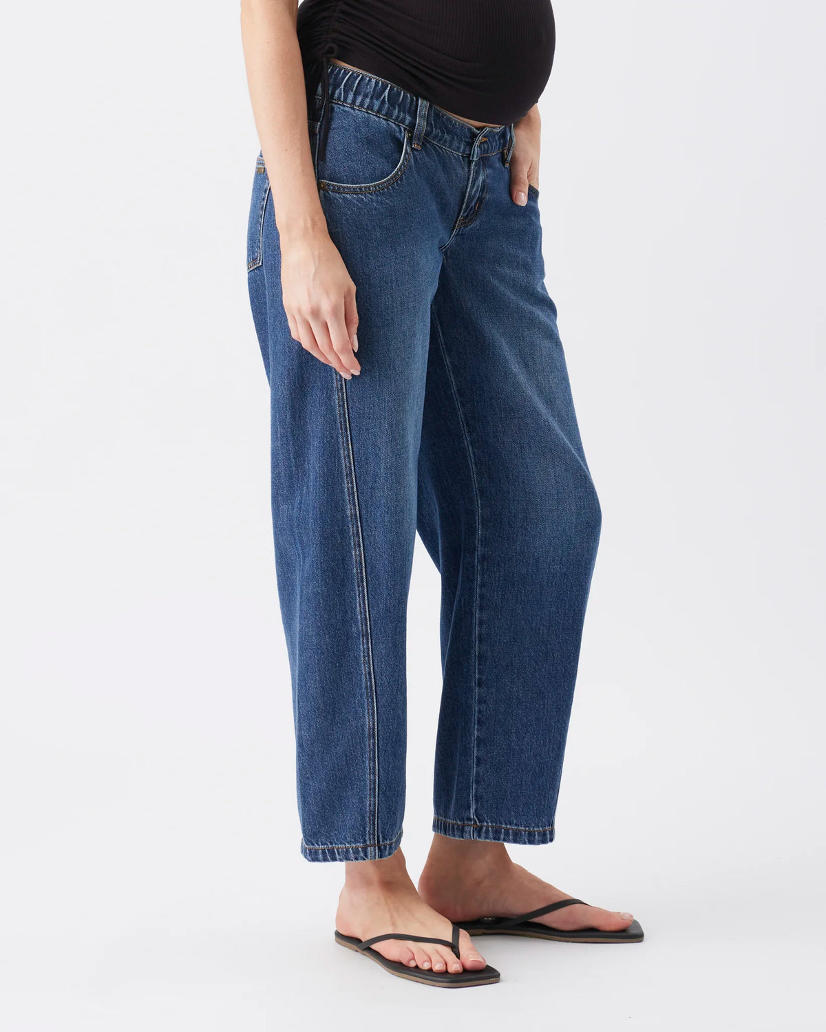 A person wearing loose-fit blue denim jeans with a straight leg and cropped hem, paired with a black top and black flip-flop sandals, photographed against a plain light gray studio background.
