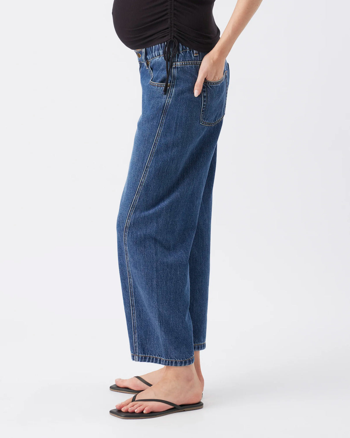 A side-profile view of a model wearing a pair of wide-leg cropped blue denim jeans with a black top and black flip-flops against a plain white studio background.
