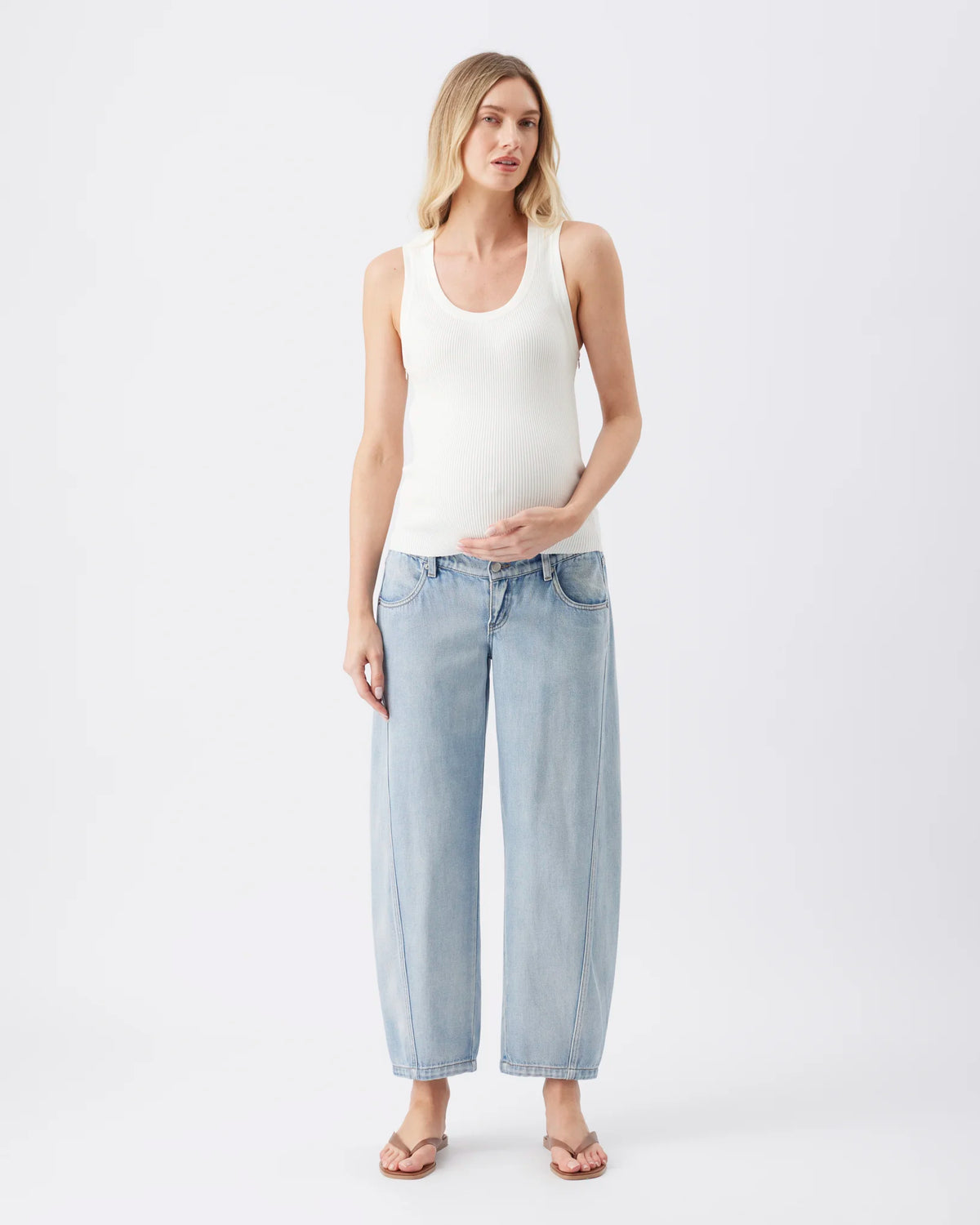 A pregnant woman stands against a light gray studio background wearing a white ribbed tank top, light-wash wide-leg cropped jeans, and tan sandals.