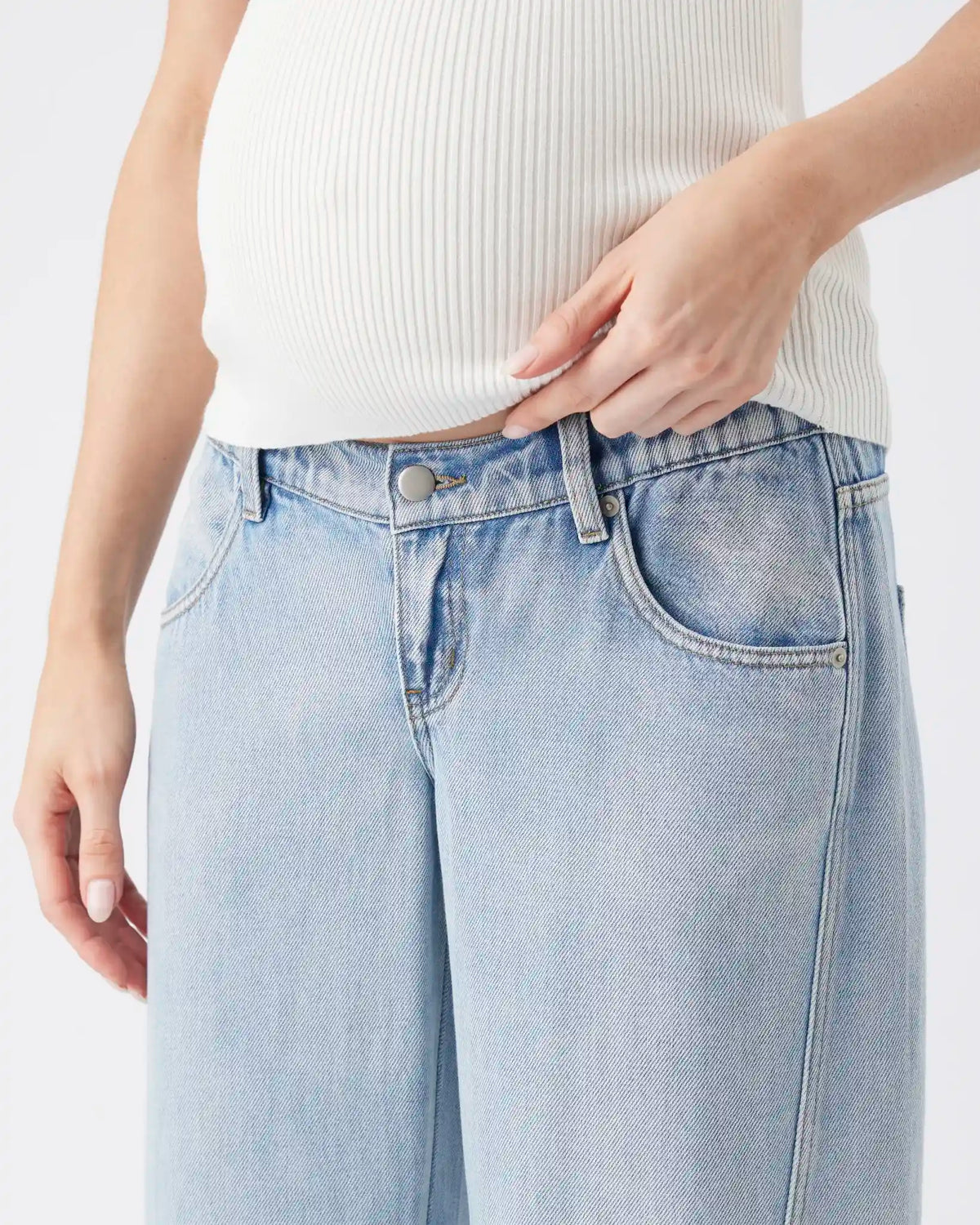 Close-up of a person wearing light-wash blue jeans and a white ribbed top, with the hand adjusting the waistband.