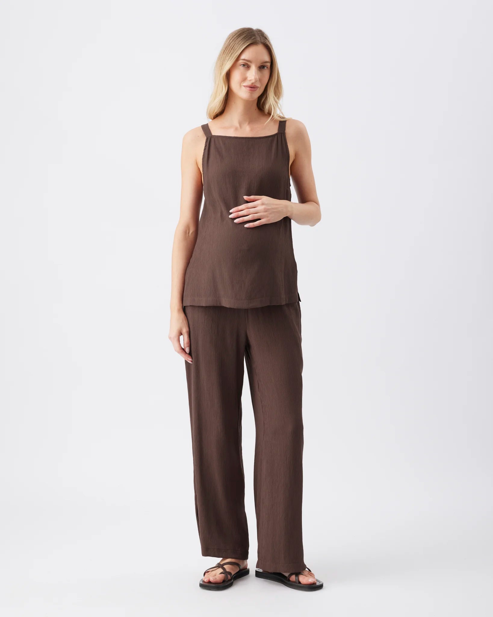 A woman modeling a matching dark brown two-piece lounge set consisting of a sleeveless top with a square neckline and wide-leg pants, paired with dark brown sandals, against a plain white studio background.