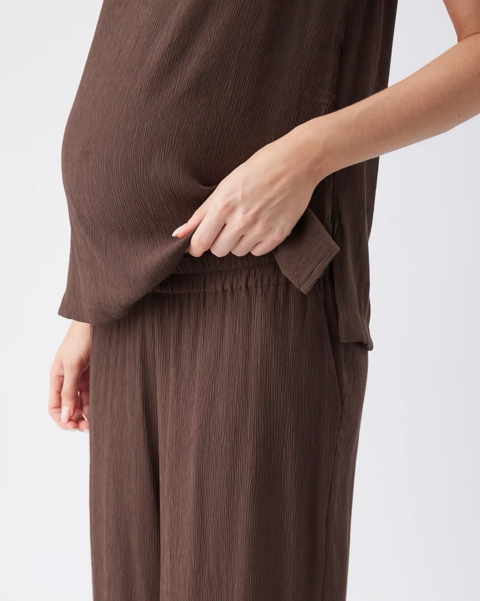 A close-up of a model wearing a dark brown crinkled two-piece outfit, featuring a sleeveless top with a side tab detail and matching pleated trousers, against a plain light background.