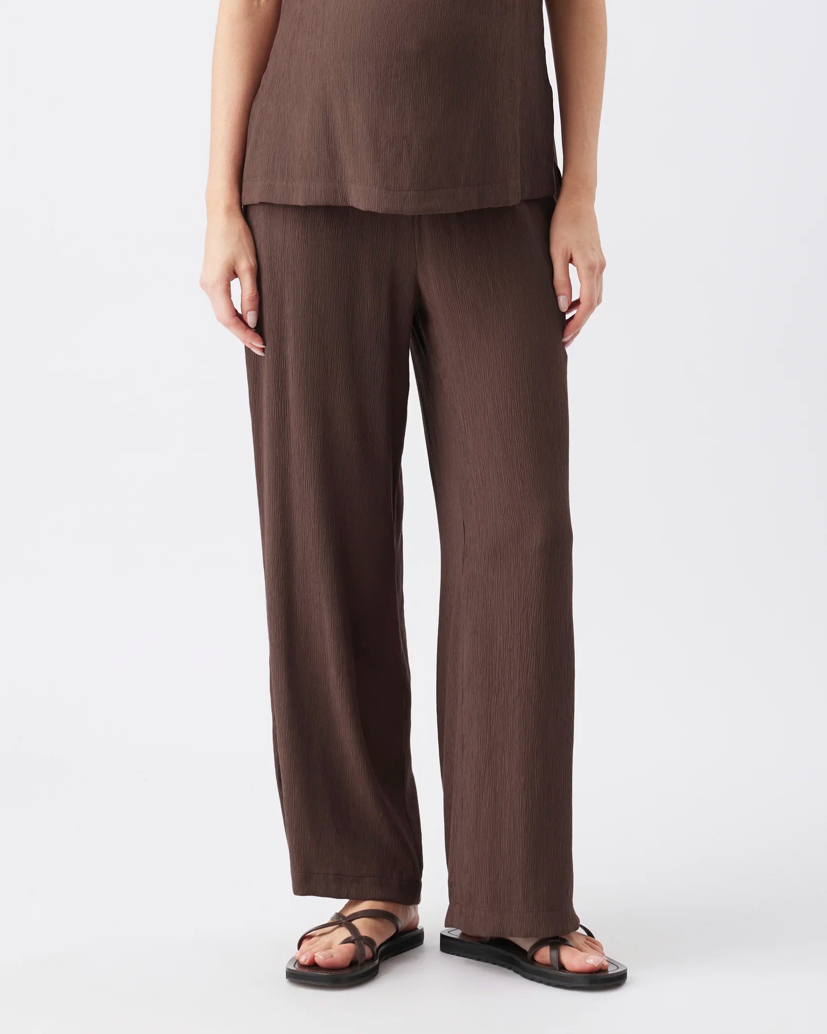 A model wearing a dark brown linen-blend top and matching wide-leg trousers, paired with black strappy sandals, shown from the waist down against a light gray studio background.