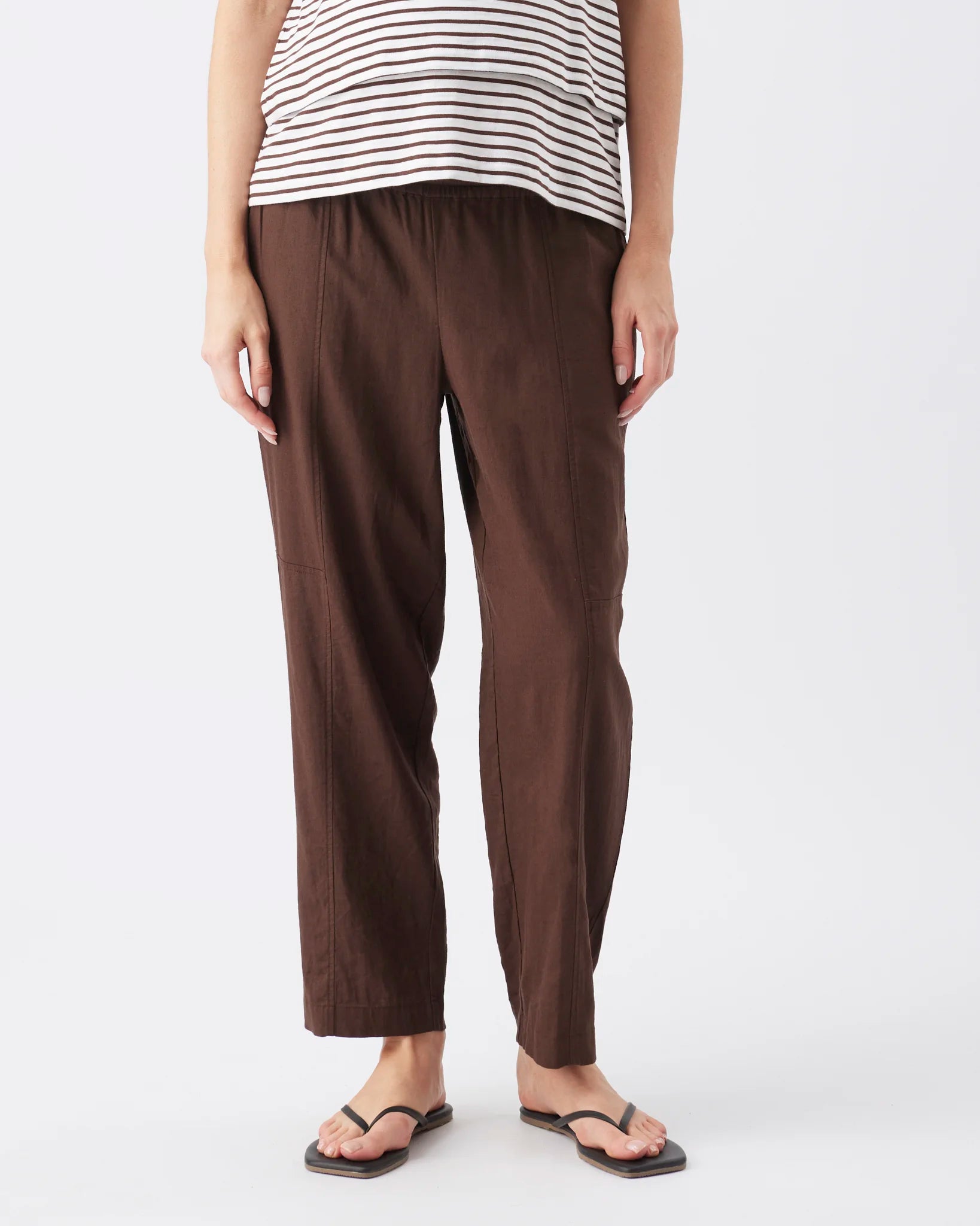 A person wearing dark brown cropped pants with a striped top and black flip-flop sandals, shown from the waist down against a plain light gray studio background.
