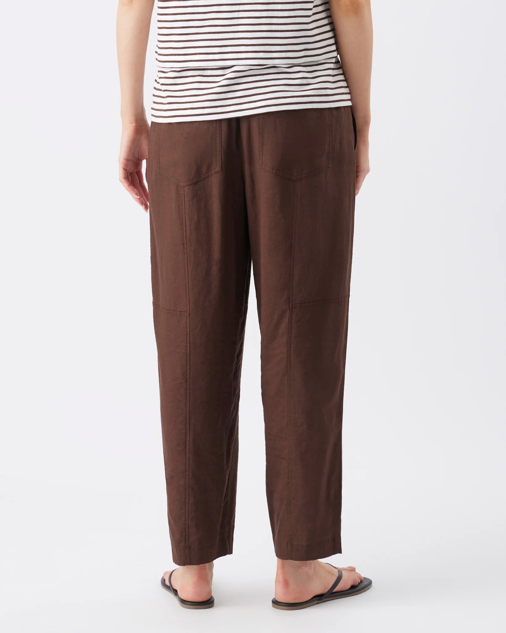 A person shown from the back wearing loose brown pants, a white top with thin black horizontal stripes, and black sandals against a plain light gray studio background.