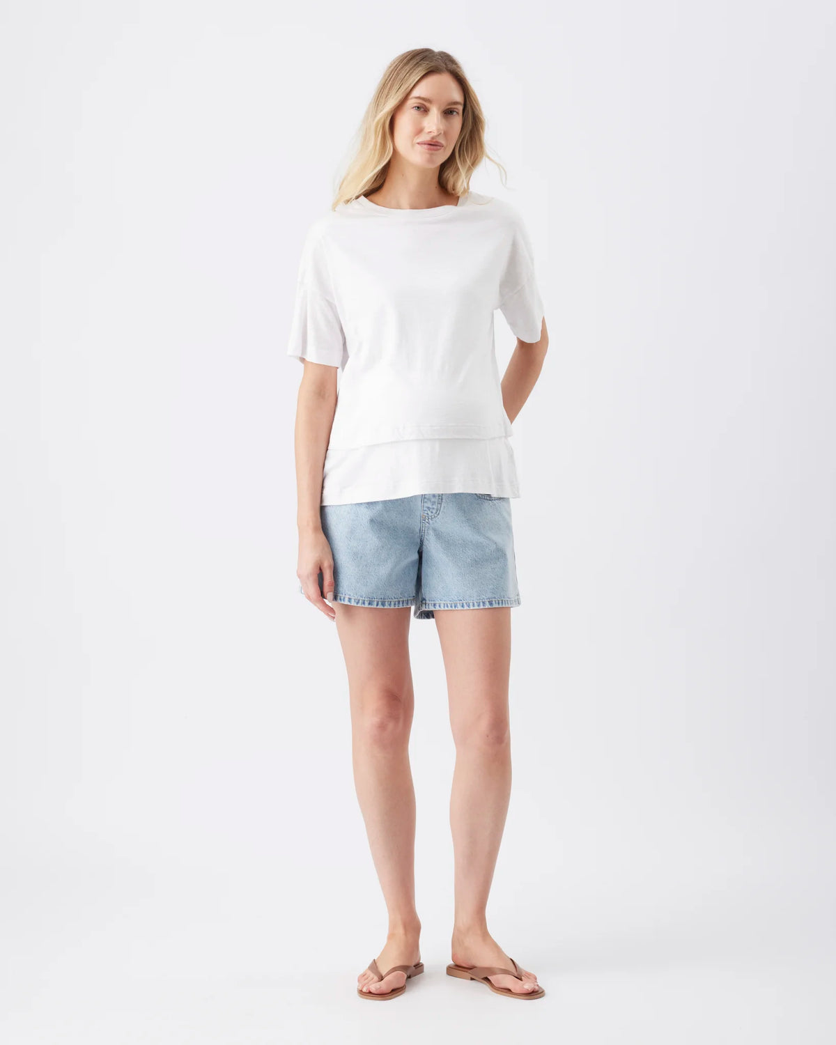 A woman modeling a white short-sleeve crewneck top and light-wash denim shorts against a plain white studio background.