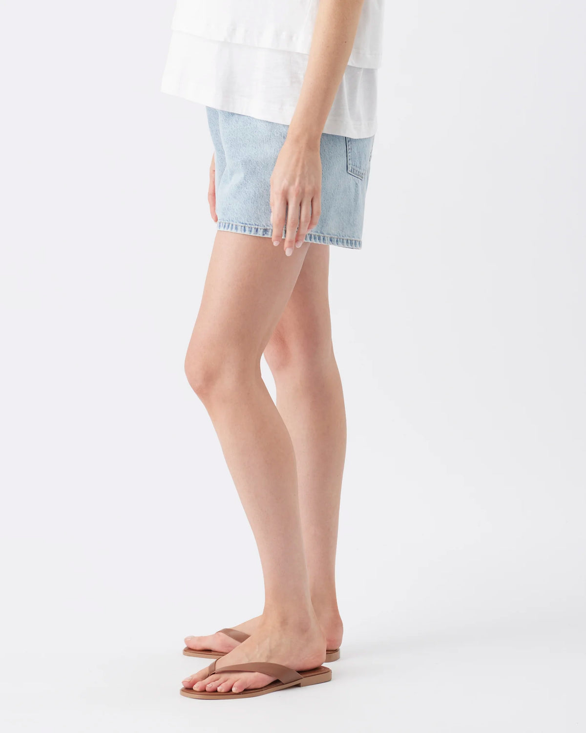 A model wearing light-wash denim shorts, a white top, and brown flip-flop sandals, shown from the waist down against a plain white studio background.