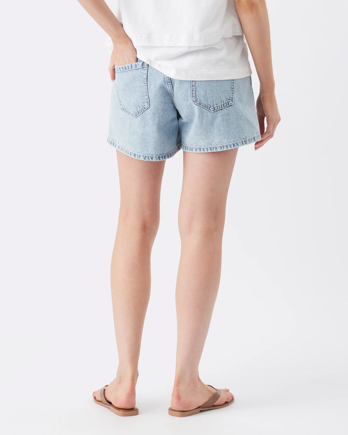 A person shown from the back wearing light-wash denim shorts with a white top and tan flip-flop sandals, photographed against a clean light gray studio background.