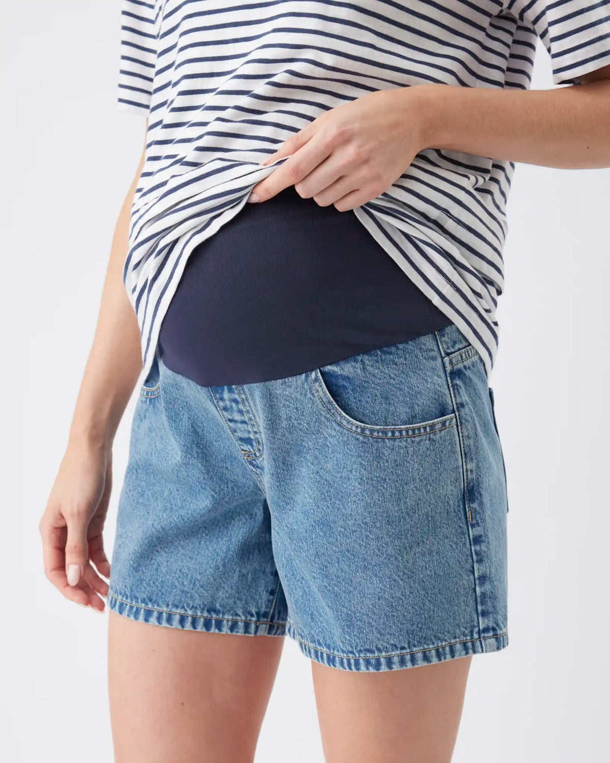 A close-up of a pregnant model wearing light-wash blue denim shorts and a navy blue maternity belly band under a navy and white striped t-shirt, against a plain white background.