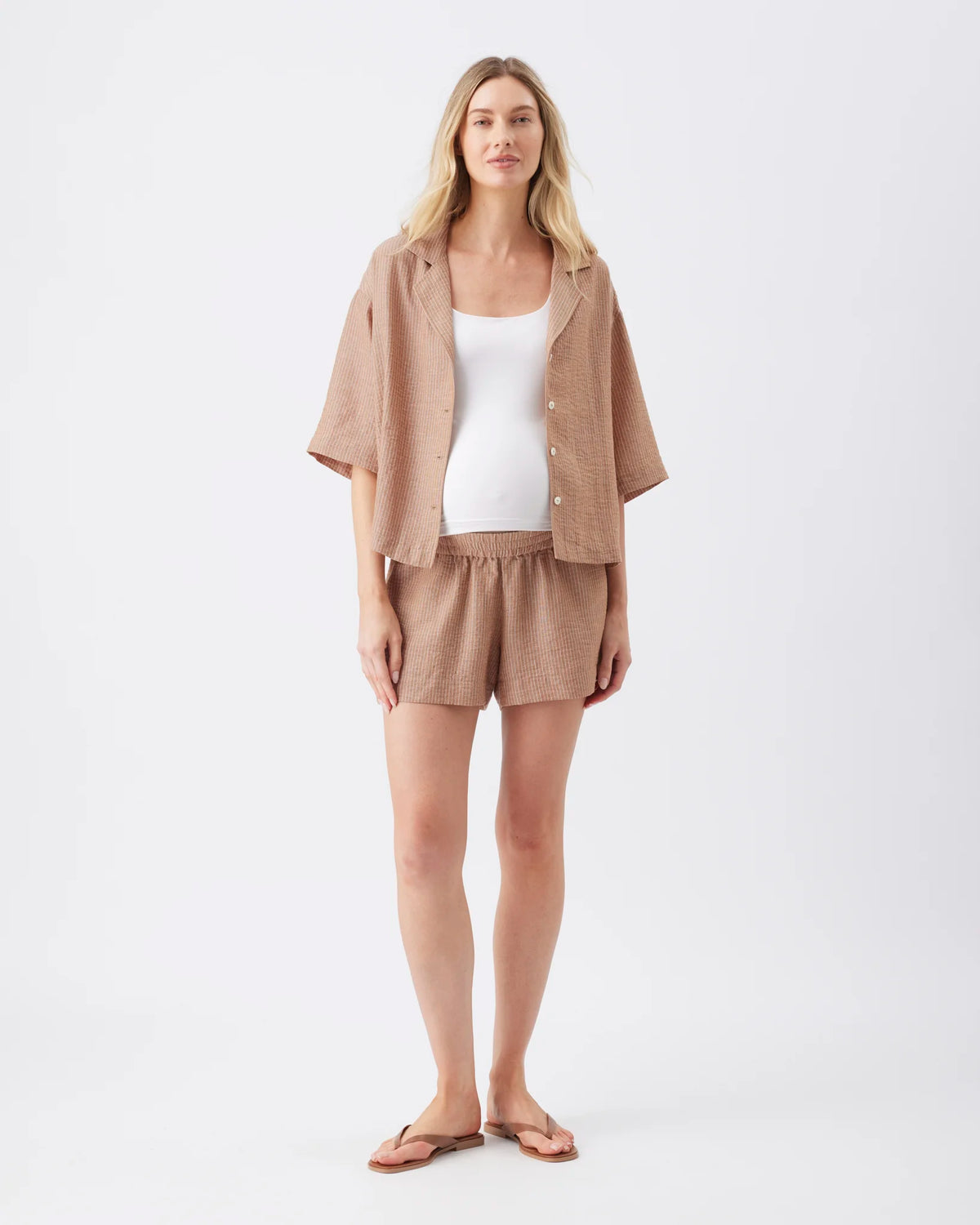 A woman modeling a light brown linen lounge set consisting of a short-sleeve button-up shirt and matching shorts, worn over a white tank top and finished with brown sandals, on a white studio background.