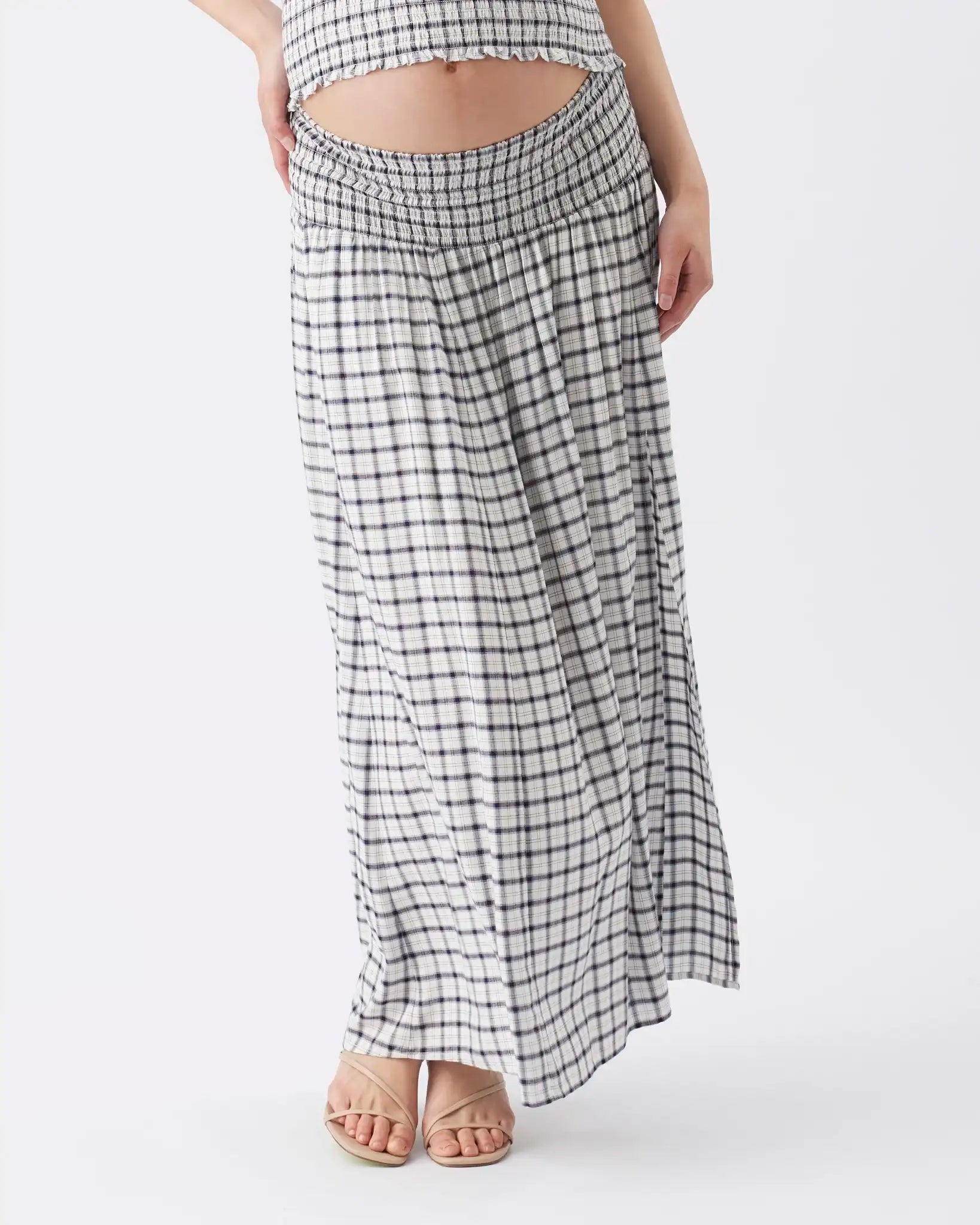 A model wearing a black and white checkered maxi skirt with a smocked waistband, paired with neutral-colored flat sandals, against a plain light gray background.