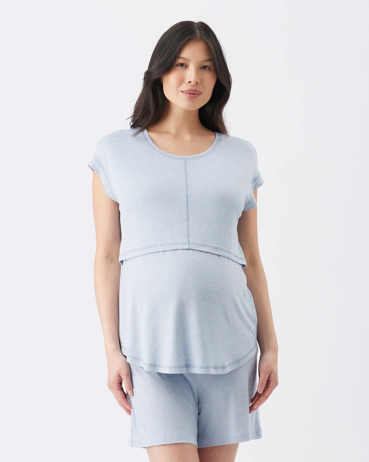 A woman modeling a light blue maternity sleep set consisting of a short-sleeve crewneck top and matching shorts against a plain white studio background.