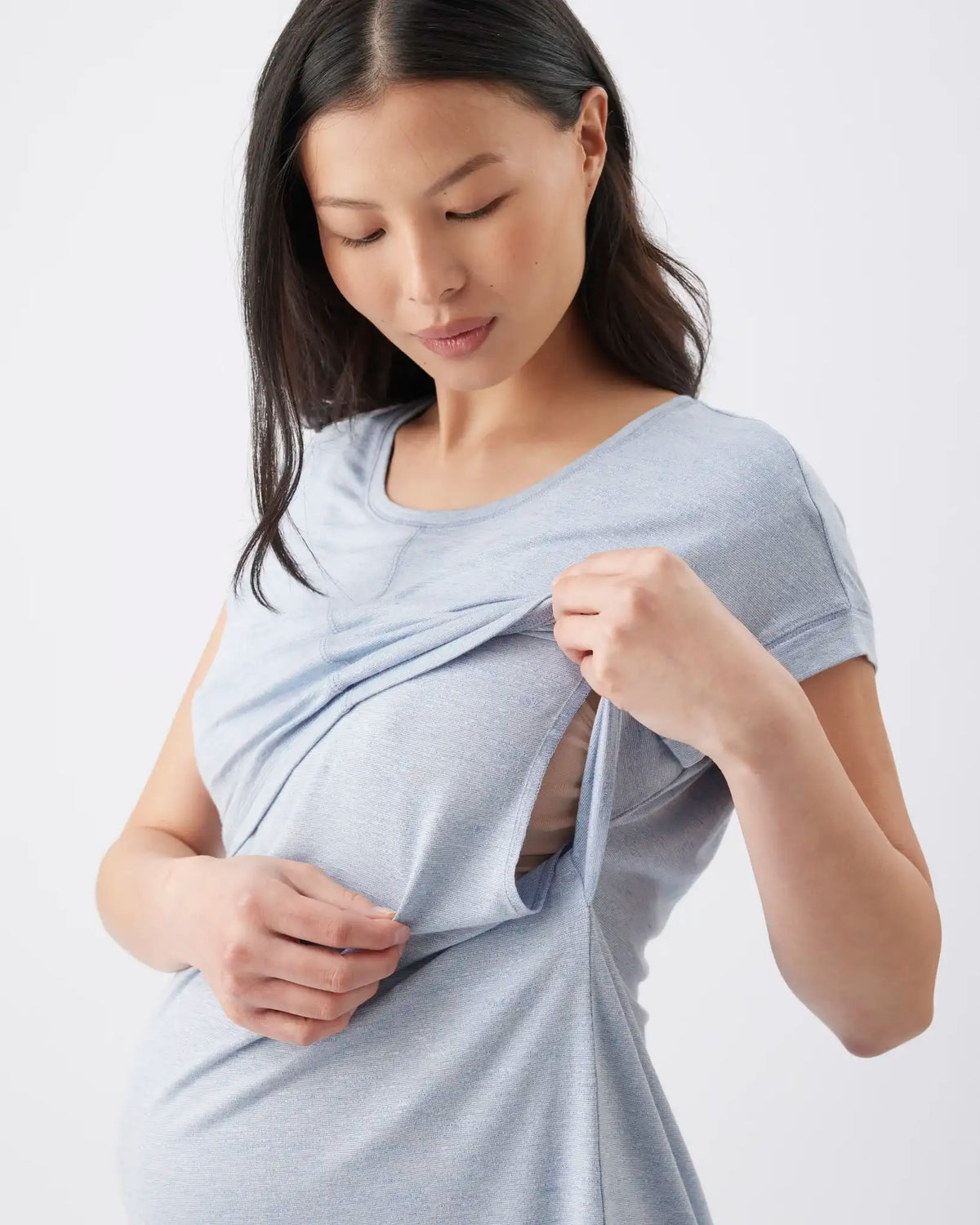 A pregnant woman wearing a light gray maternity tank top, lifting the front fabric to show a built-in pocket on the opposite side of the chest.