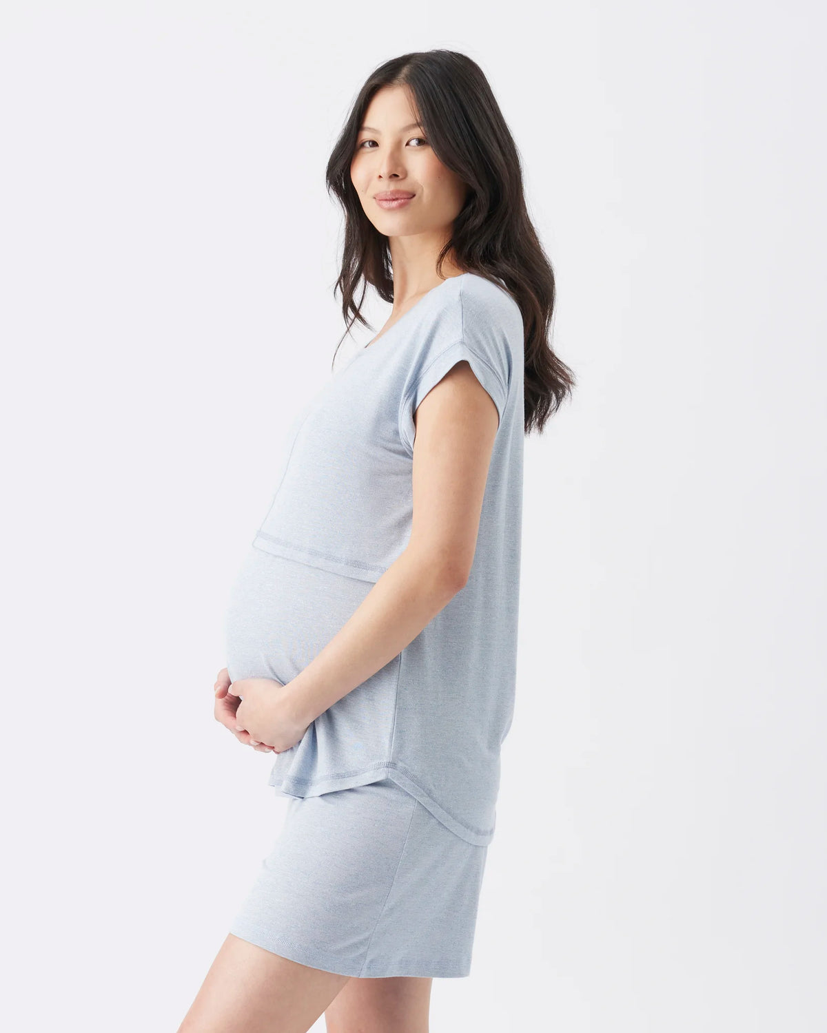 A pregnant woman with long dark hair is shown in profile wearing a light blue maternity top and matching mini skirt against a plain white studio background.