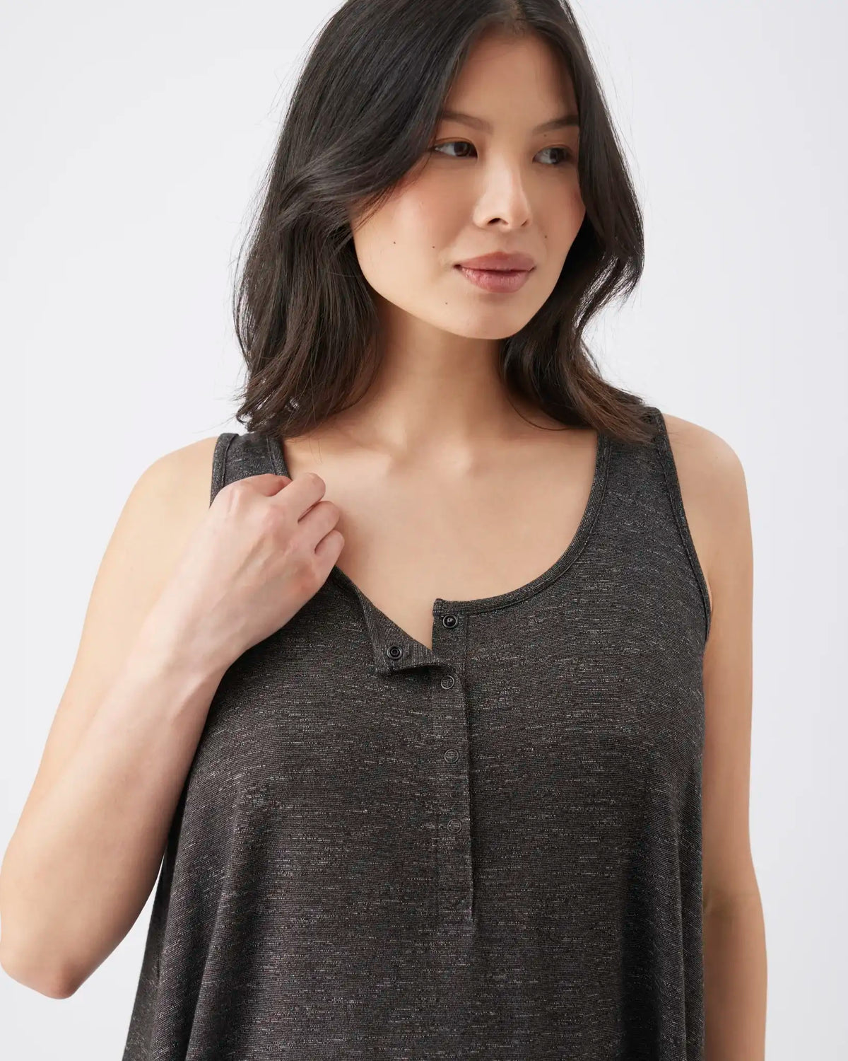 A woman wearing a dark gray heathered sleeveless henley tank top, holding the shoulder strap with one hand against a plain light gray background.