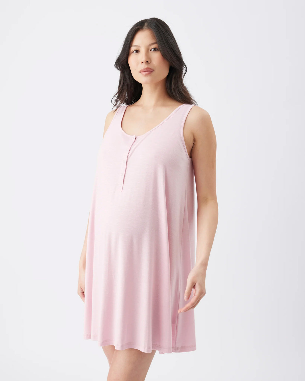 A pregnant woman wearing a light pink sleeveless maternity nightgown, standing against a plain white studio background.