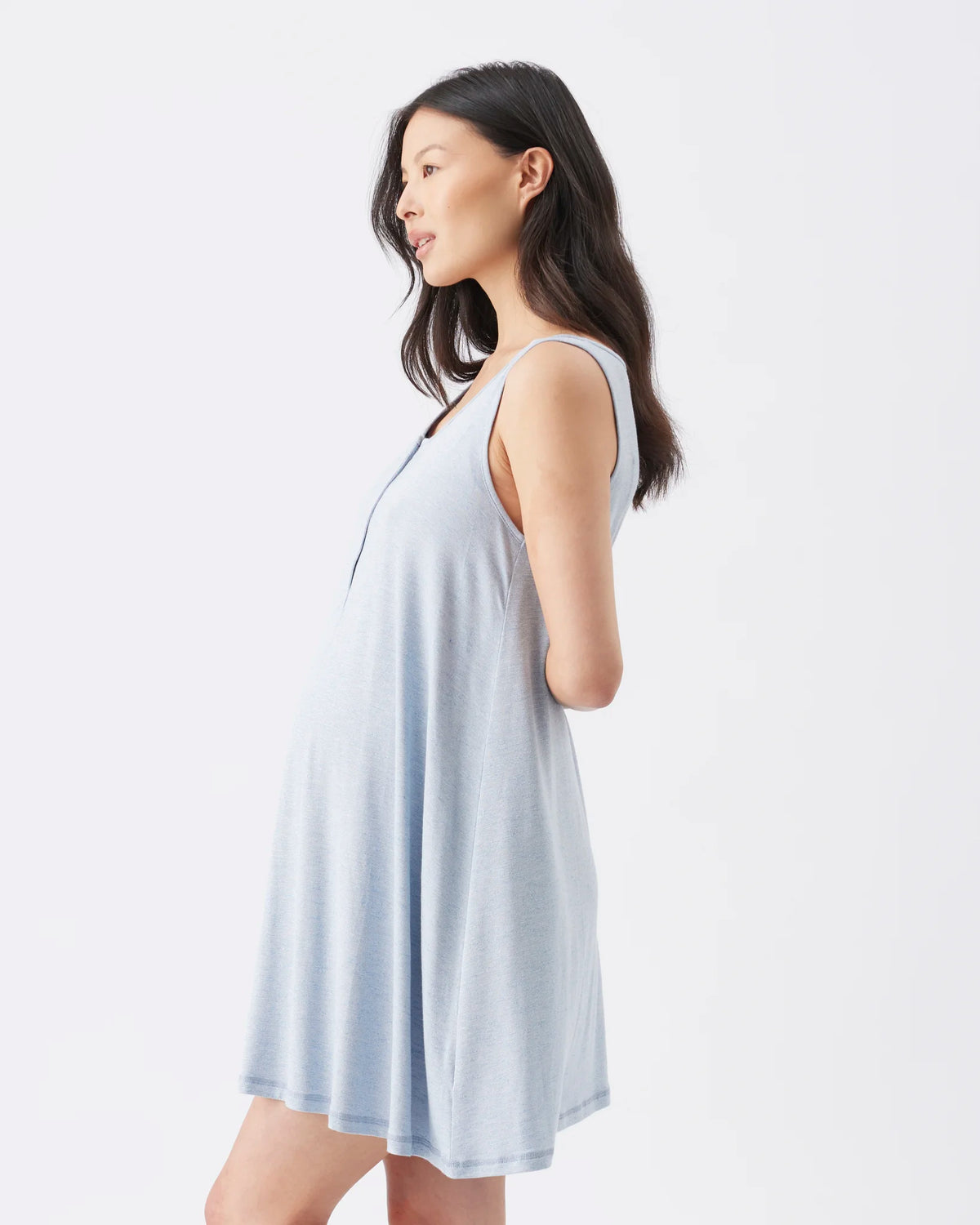 A woman wearing a light blue sleeveless maternity nightgown or shift dress, shown in profile against a clean white studio background.