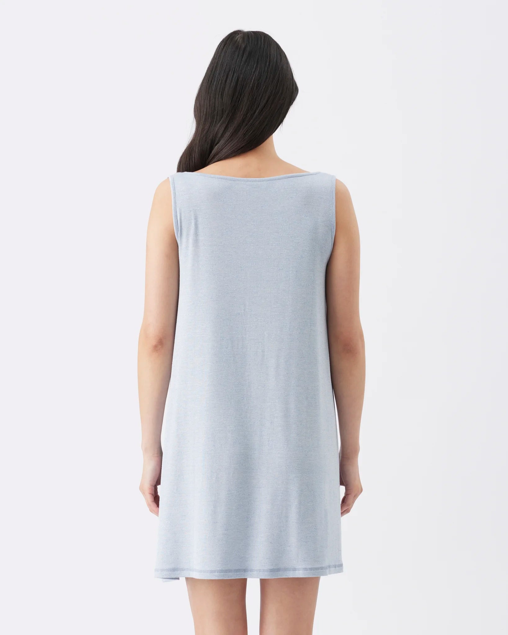 A woman viewed from the back wearing a sleeveless, knee-length light gray tunic or nightshirt against a plain white studio background.