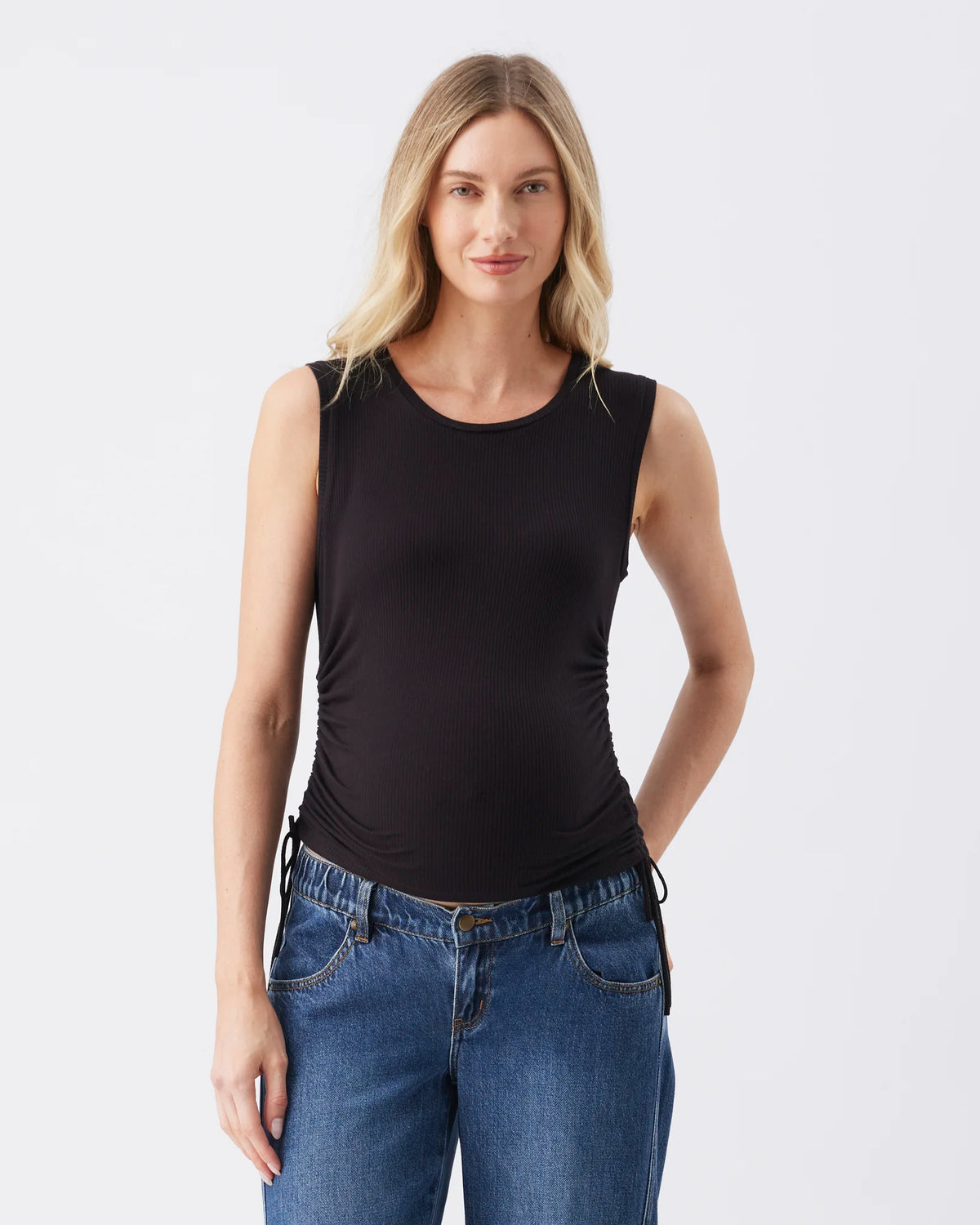 A woman modeling a black sleeveless scoop-neck tank top with ruched detailing at the sides, paired with blue jeans, against a plain light gray studio background.