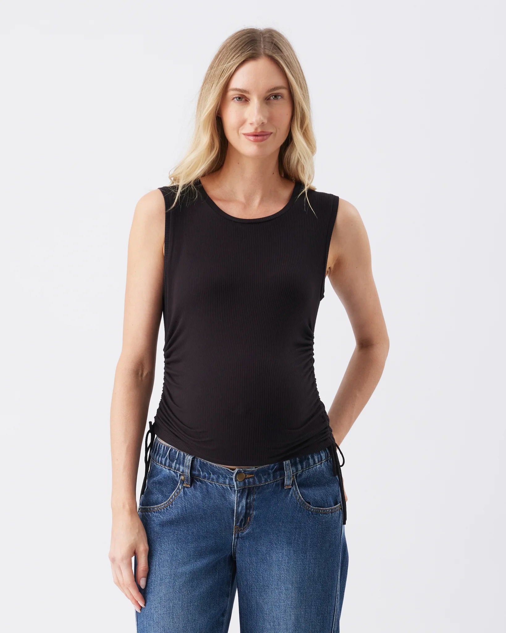 A woman modeling a black sleeveless scoop-neck tank top with ruched detailing at the sides, paired with blue jeans, against a plain light gray studio background.
