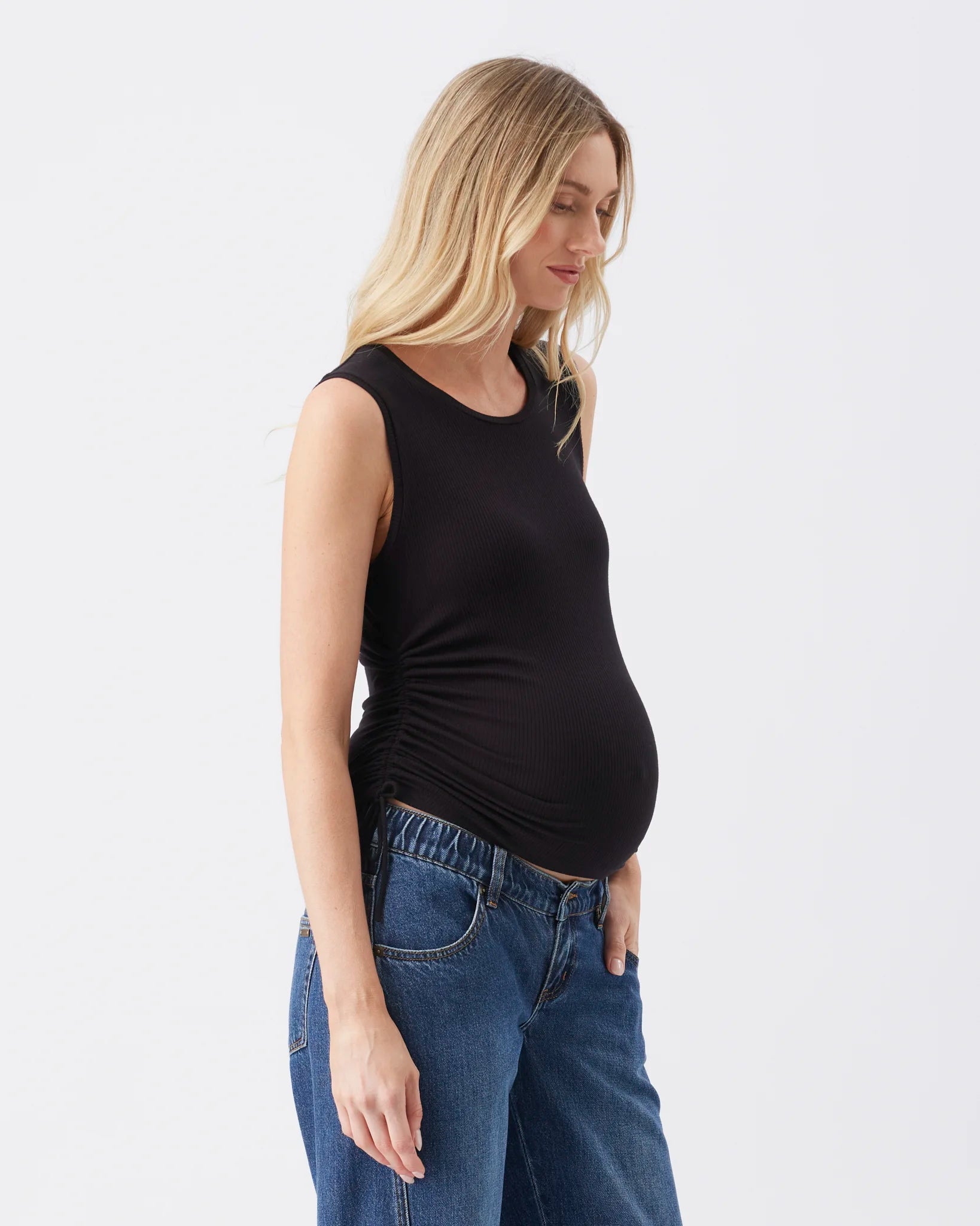 A pregnant woman wearing a black maternity tank top and blue jeans, shown in profile against a plain white studio background.