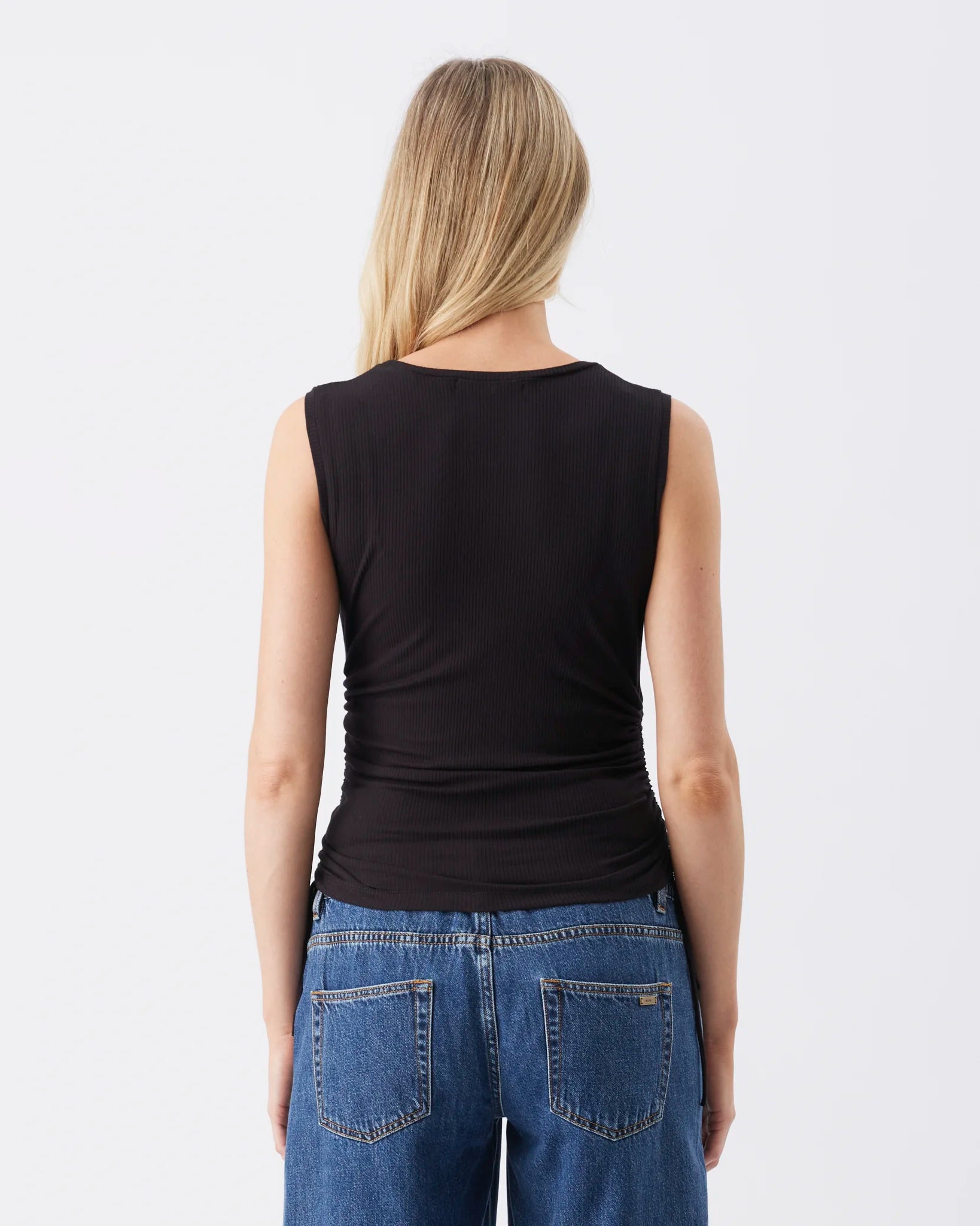 A woman viewed from the back wearing a black sleeveless top and blue jeans against a plain light gray studio background.