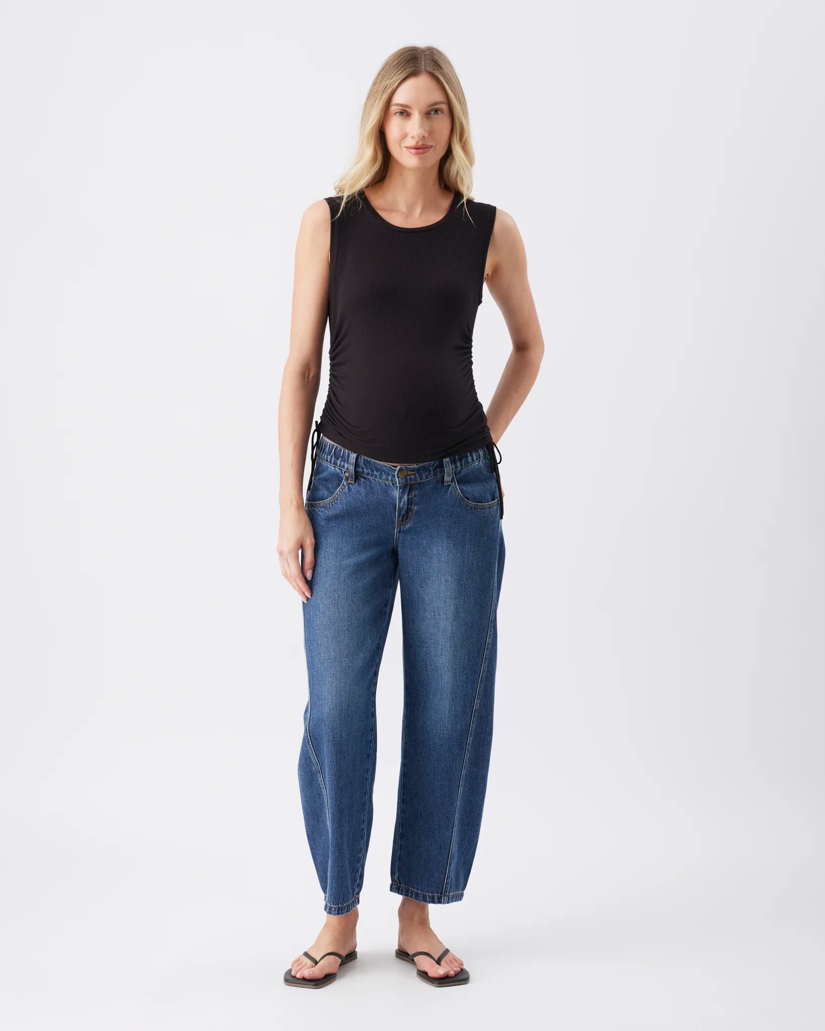 A woman modeling a black sleeveless top and blue wide-leg cropped jeans with dark sandals, standing against a plain light gray studio background.