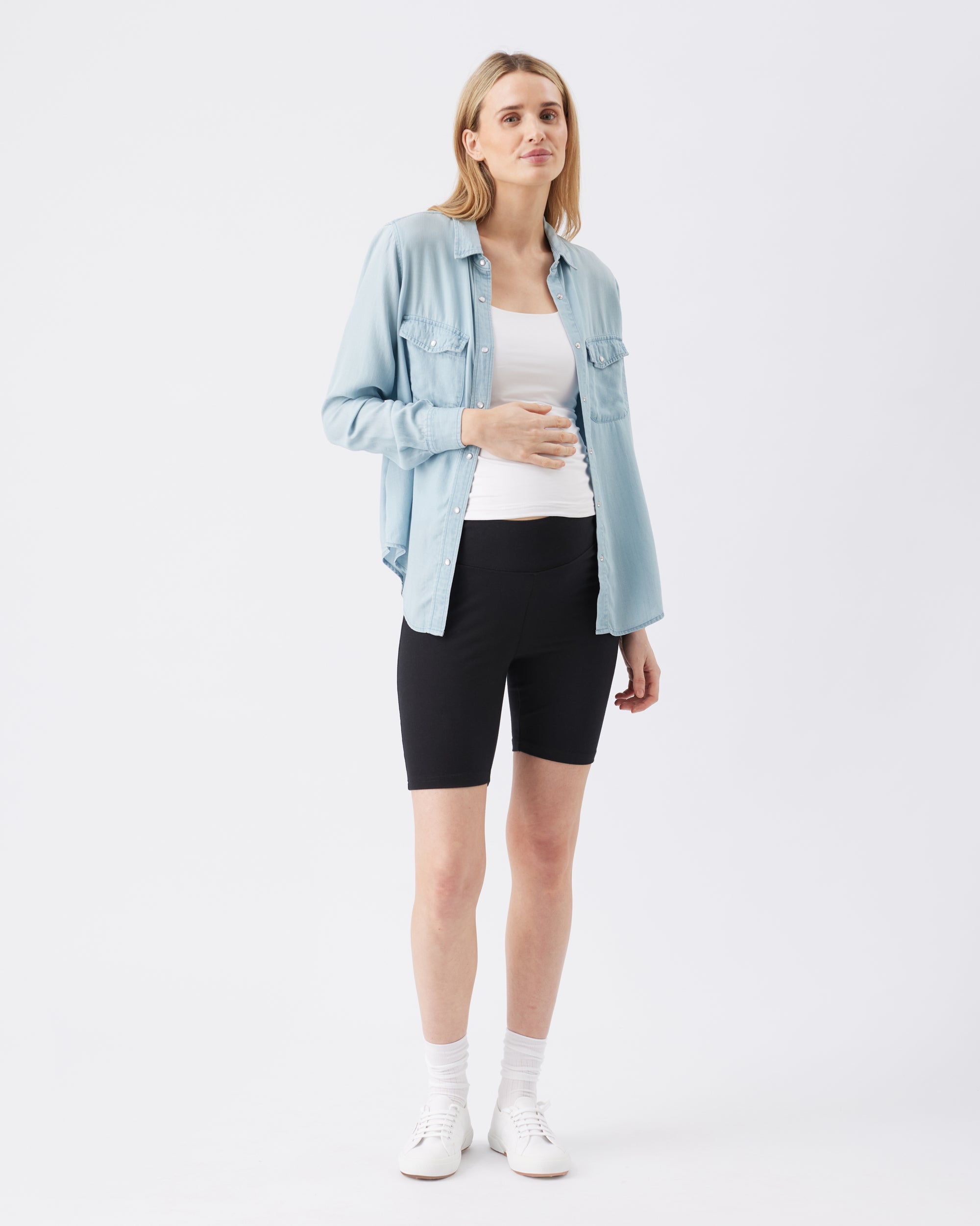 Woman wearing a light blue denim jacket, white top, and black active shorts on a white background