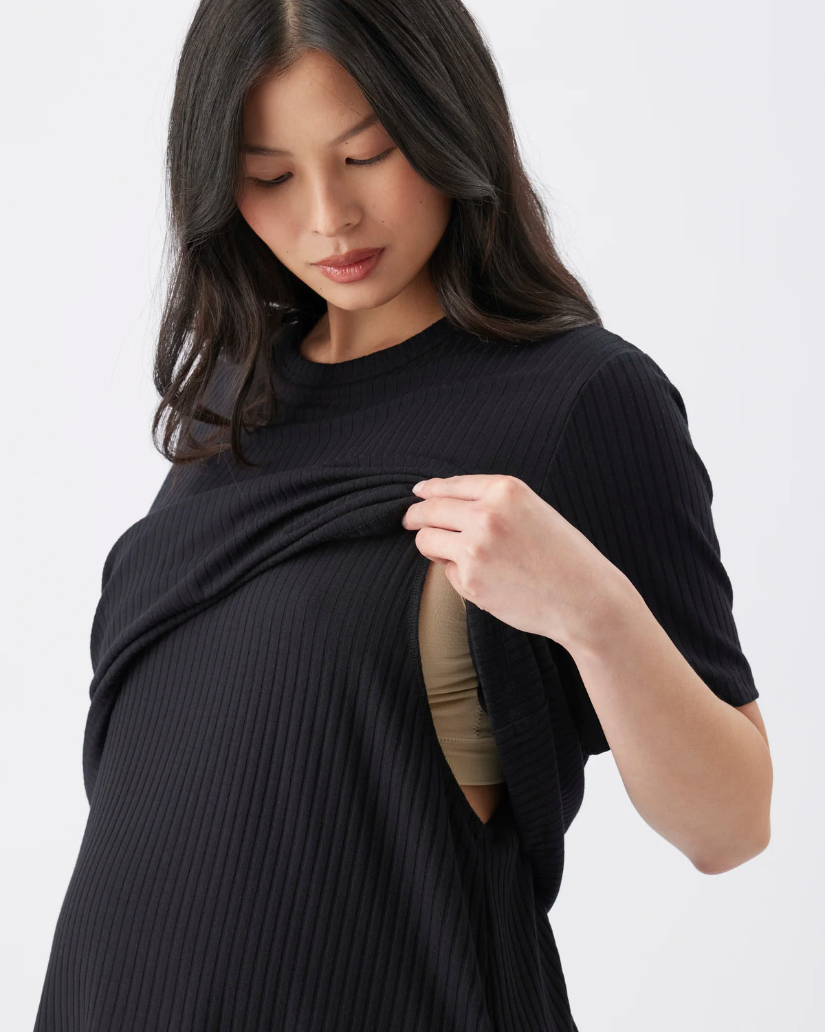 A pregnant woman wearing a black ribbed short-sleeve maternity top, lifting the fabric to reveal a beige/nude undergarment underneath, set against a plain light gray background.