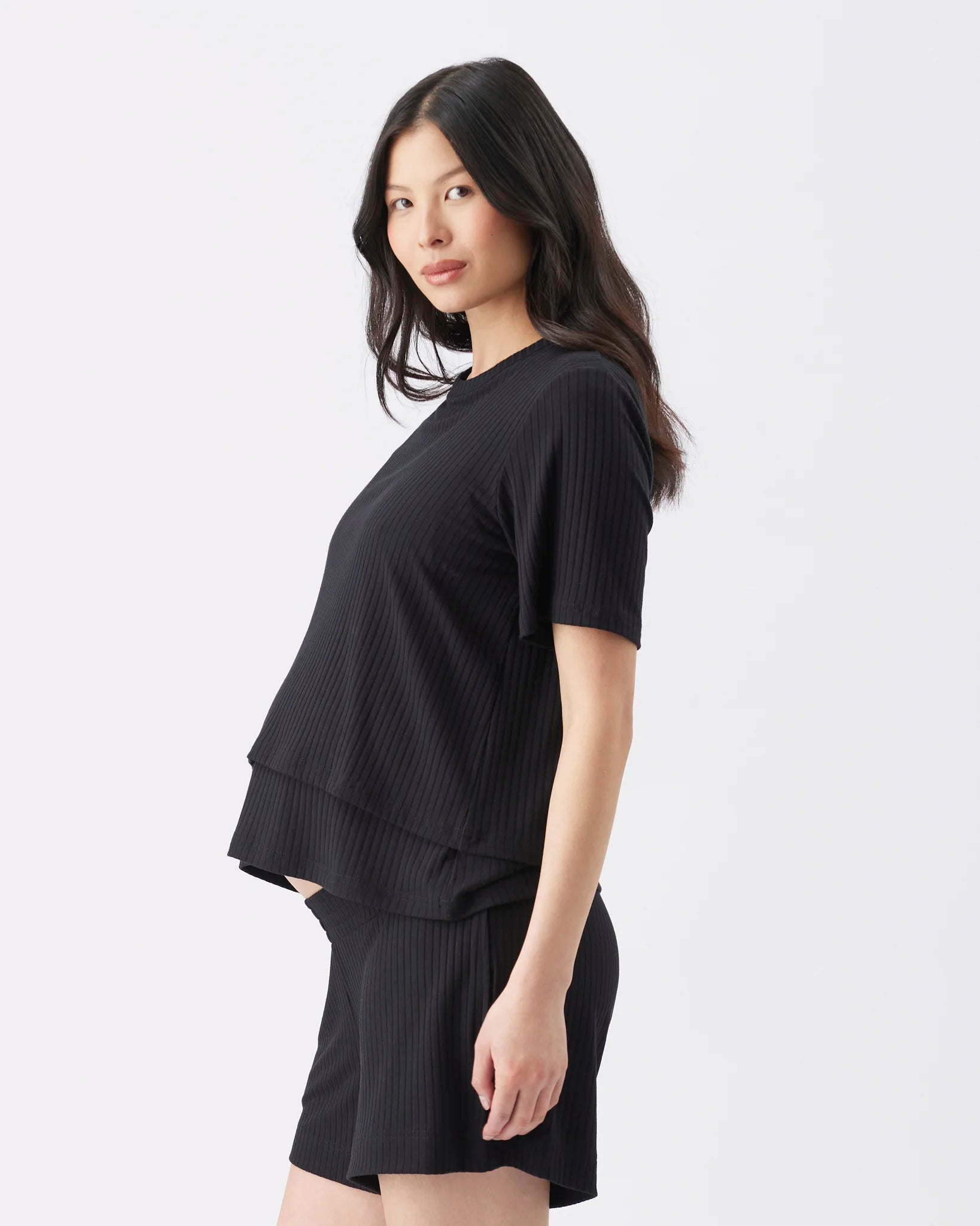 A woman modeling a black short-sleeve ribbed top and matching black shorts against a plain light gray studio background.