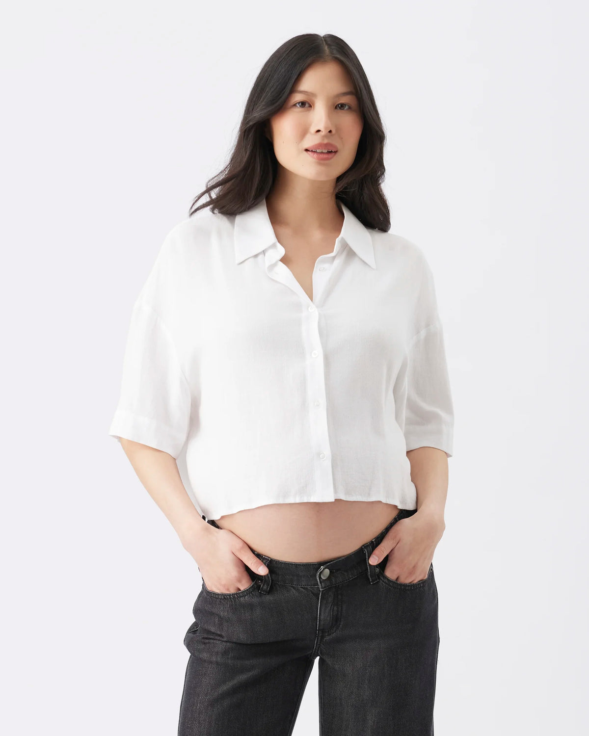 A woman modeling a white short-sleeve button-up shirt with a relaxed, cropped fit, paired with dark gray jeans, against a plain white studio background.
