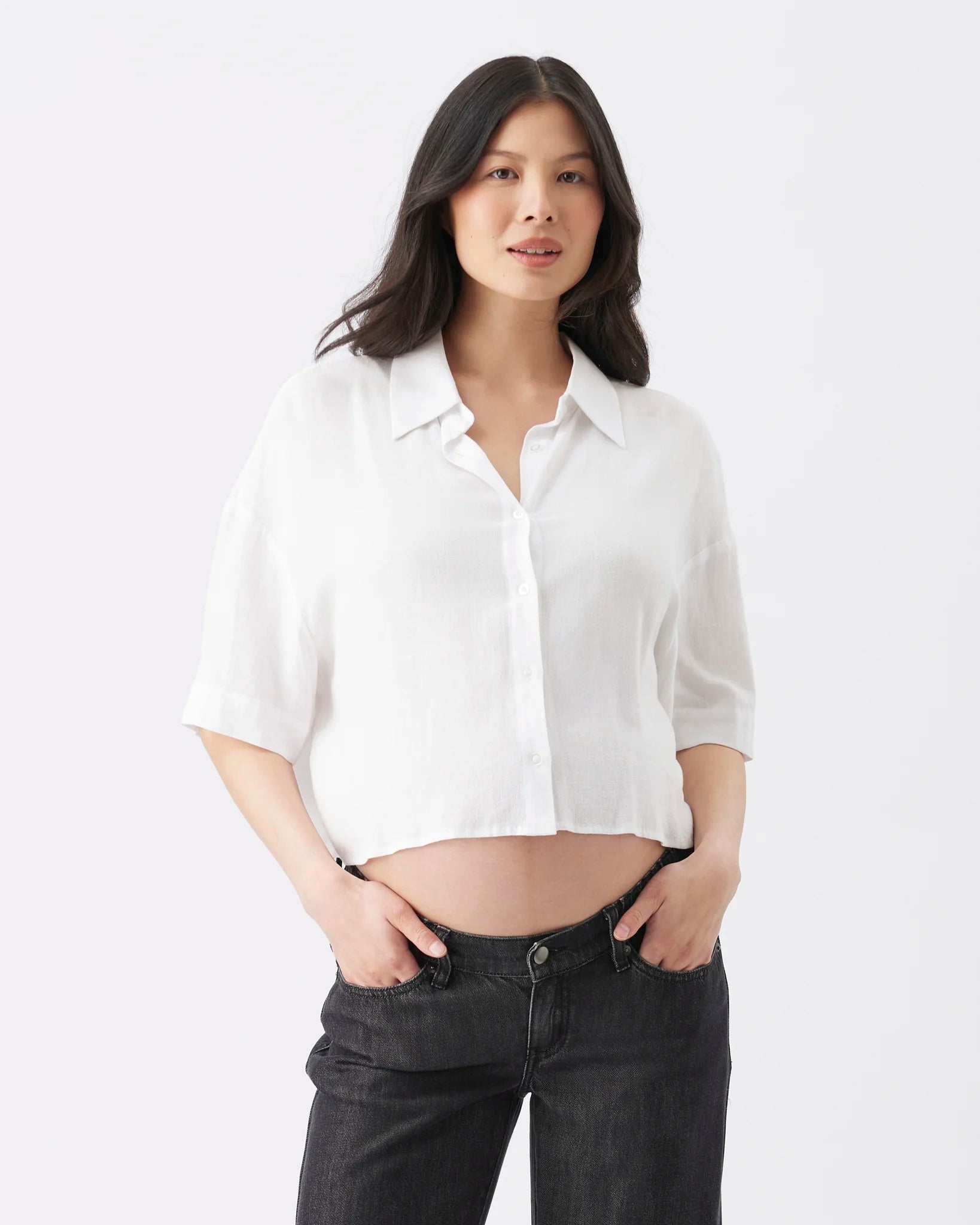 A woman modeling a white short-sleeve button-up shirt with a relaxed, cropped fit, paired with dark gray jeans, against a plain white studio background.