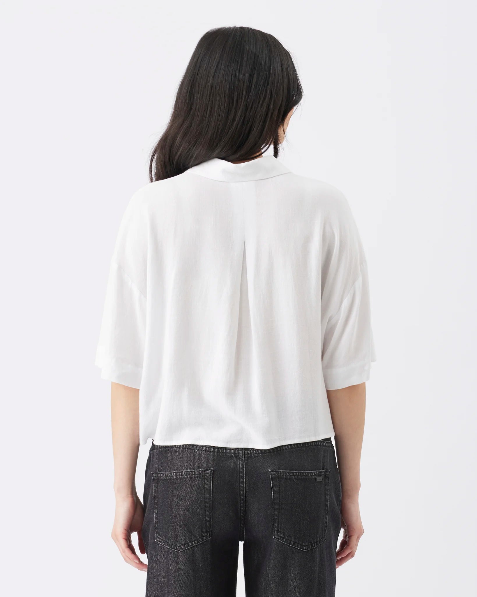 A woman viewed from behind wearing a white short-sleeve blouse with a subtle center back seam and dark gray jeans, set against a plain white studio background.