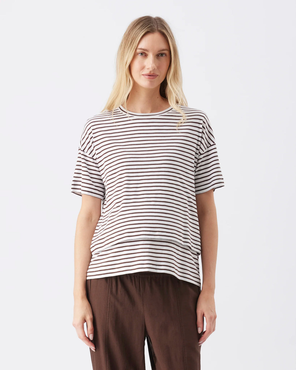 A woman modeling a short-sleeve crewneck t-shirt with thin horizontal black stripes on a white background, paired with brown pants, against a light gray studio backdrop.