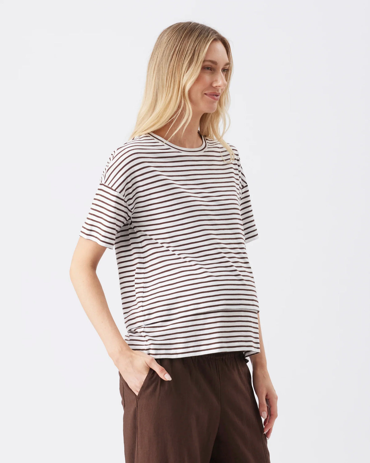 A woman wearing a short-sleeve crewneck t-shirt with thin horizontal black stripes on a white background, paired with brown pants, photographed against a light gray studio backdrop.