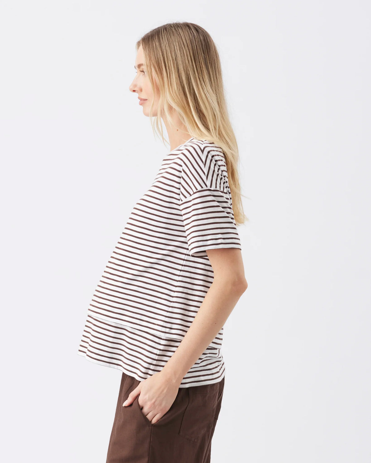 A woman in side profile wearing a short-sleeve maternity top with thin black horizontal stripes on an off-white base, paired with dark brown pants, against a plain white studio background.