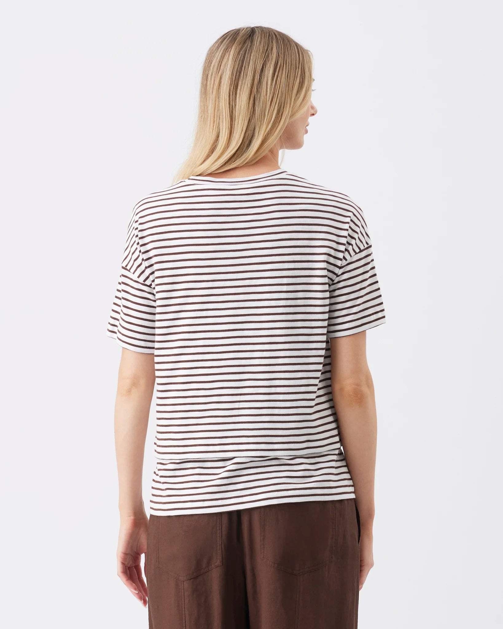 A woman shown from the back wearing a short-sleeve crewneck t-shirt with thin horizontal dark brown stripes on an off-white base, paired with brown pants, against a plain white background.