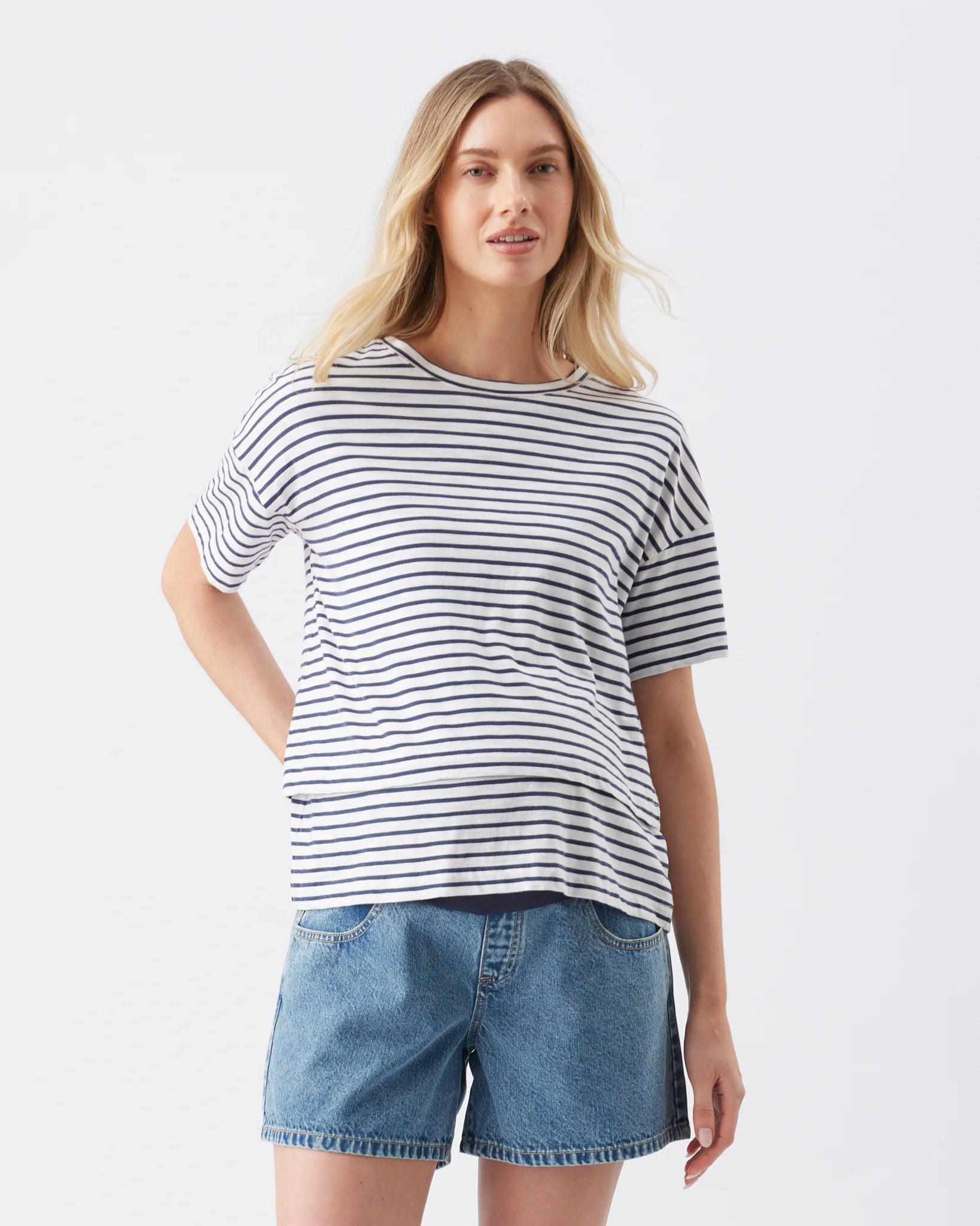 A woman modeling a short-sleeve crewneck t-shirt with thin navy and white horizontal stripes, paired with light-wash denim shorts, against a plain light gray studio background.