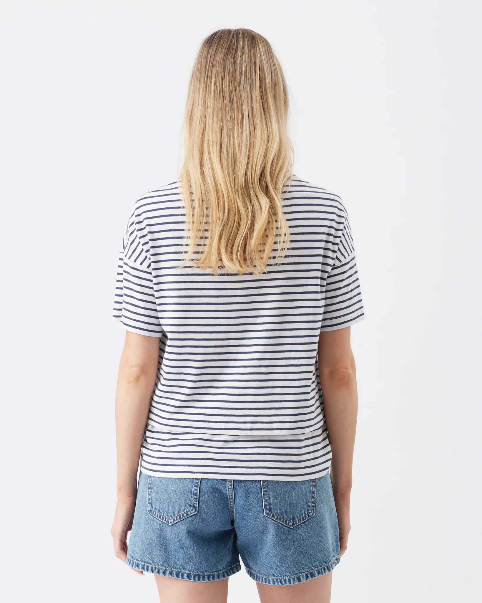 A woman viewed from the back wearing a white short-sleeve T-shirt with thin navy horizontal stripes and light blue denim shorts against a plain white studio background.
