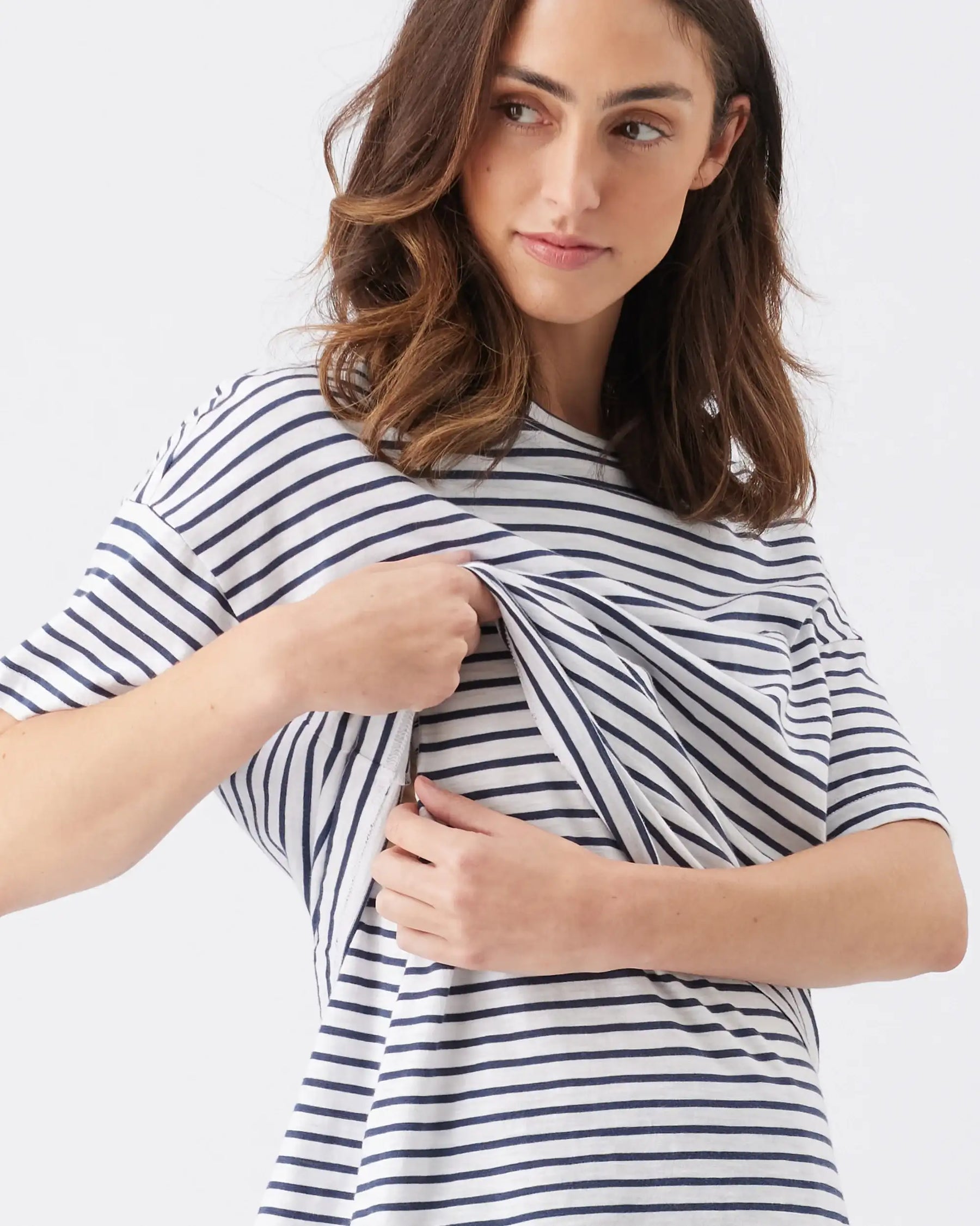 A woman wearing a navy and white horizontally striped short-sleeve top, holding the fabric at the chest to demonstrate a stretch or fit feature, against a plain light gray background.