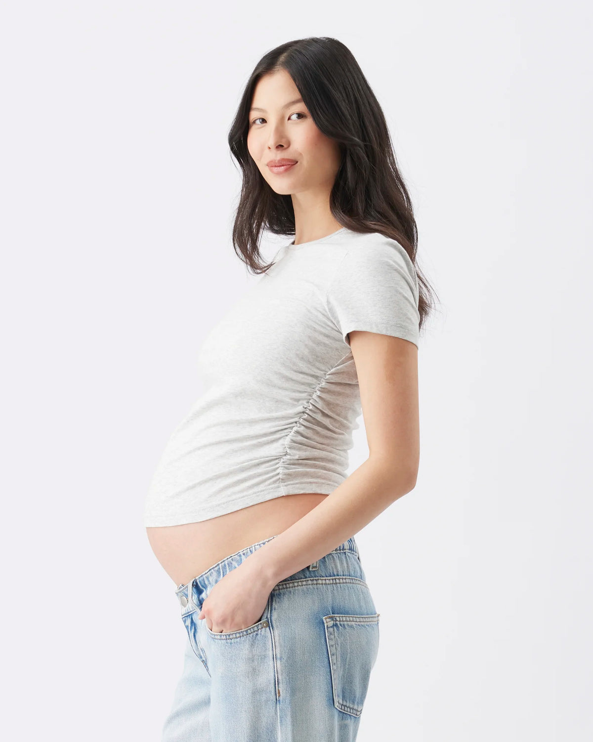 A pregnant woman wearing a white ruched short-sleeve crop top and light blue jeans, standing against a solid white studio background.