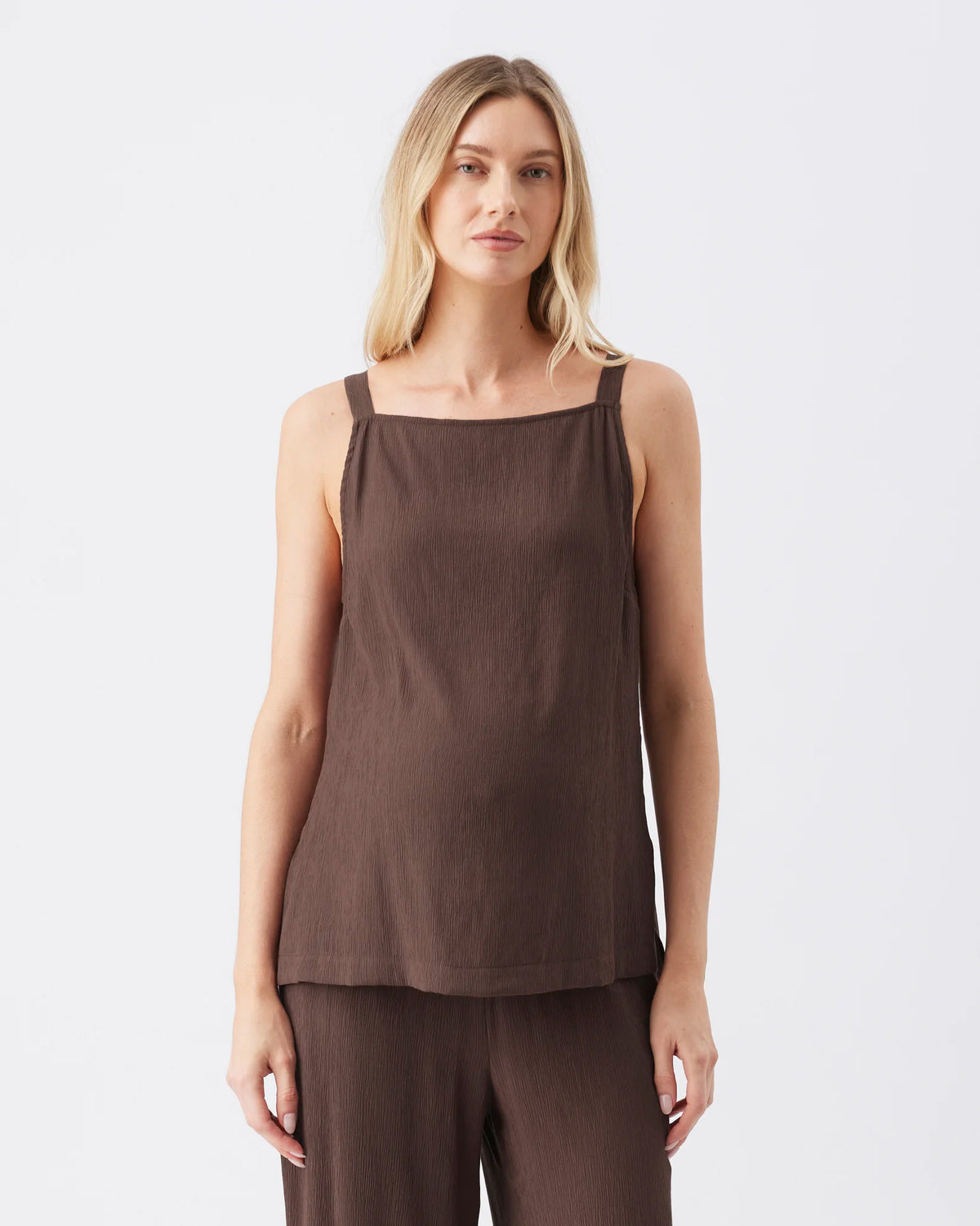 A woman modeling a sleeveless dark brown top with a square neckline and matching brown pants against a plain light gray studio background.