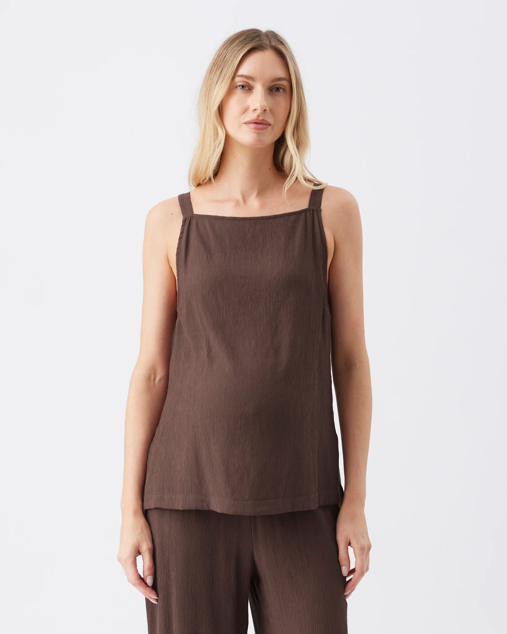 A woman modeling a sleeveless dark brown top with a square neckline and matching brown pants against a plain light gray studio background.