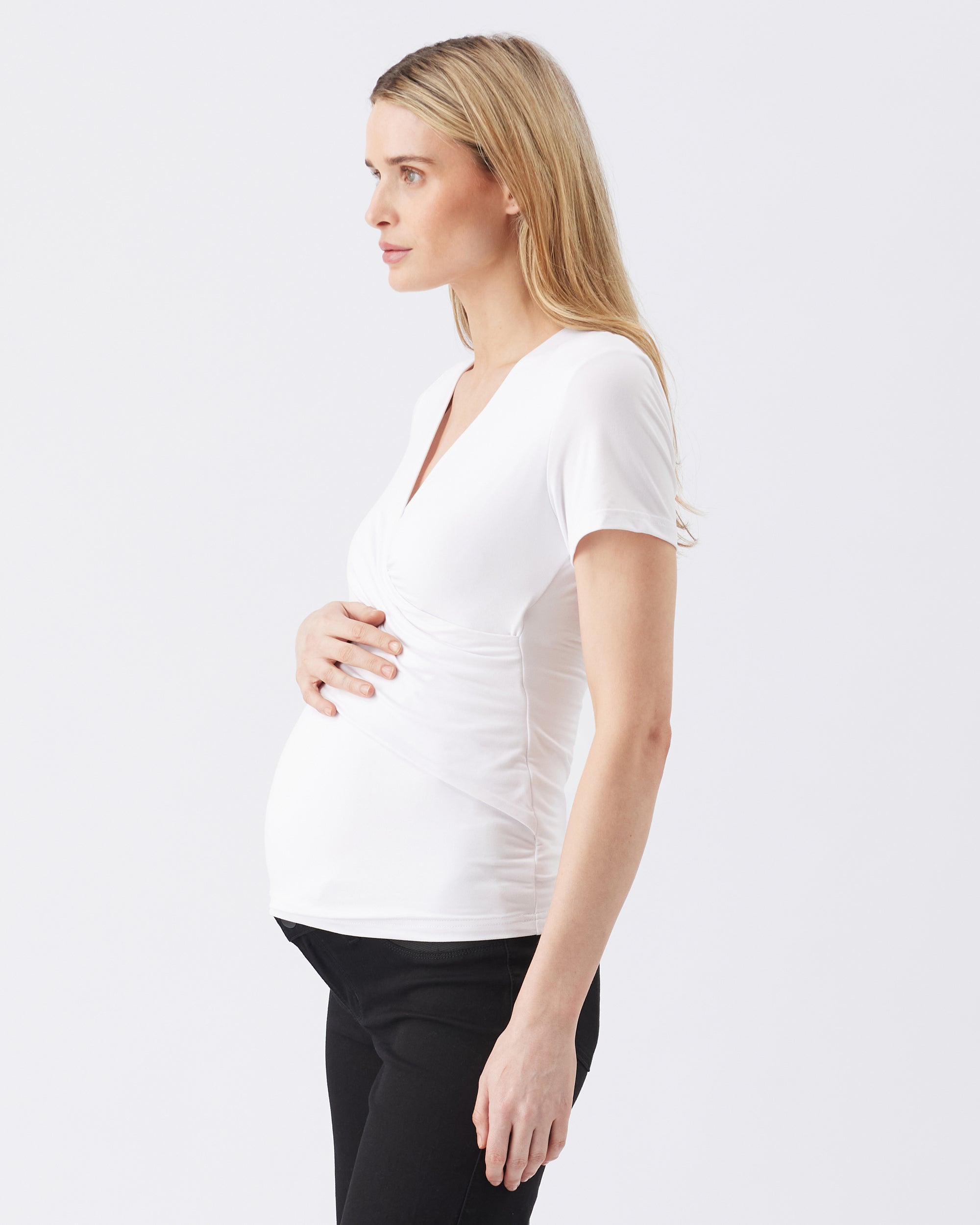 Side view of a pregnant woman wearing a white top and black pants on a plain background