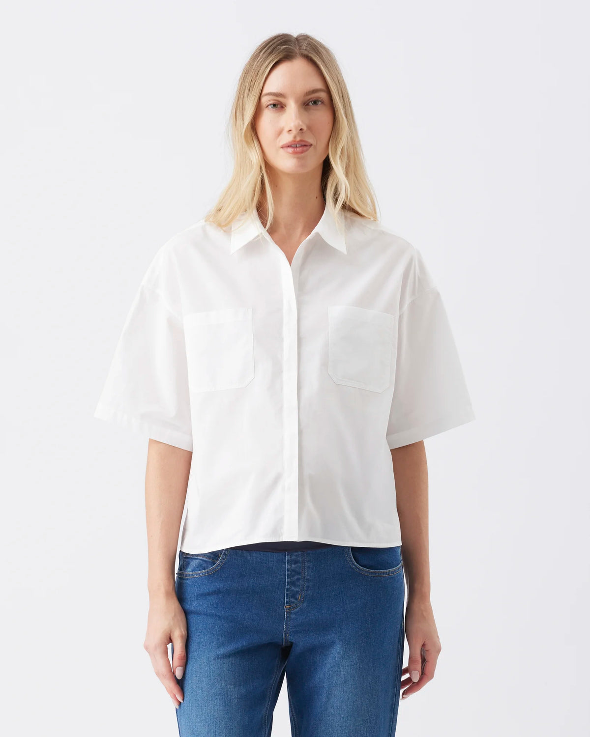 A blonde woman wearing a white short-sleeve button-up shirt with two chest pockets and blue jeans, standing against a plain light gray studio background.