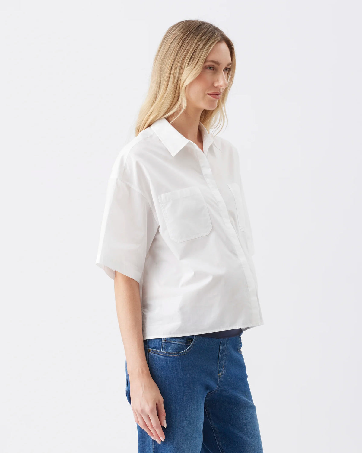 A woman wearing a white short-sleeve button-up shirt with a chest pocket, paired with blue jeans, photographed against a clean white studio background.