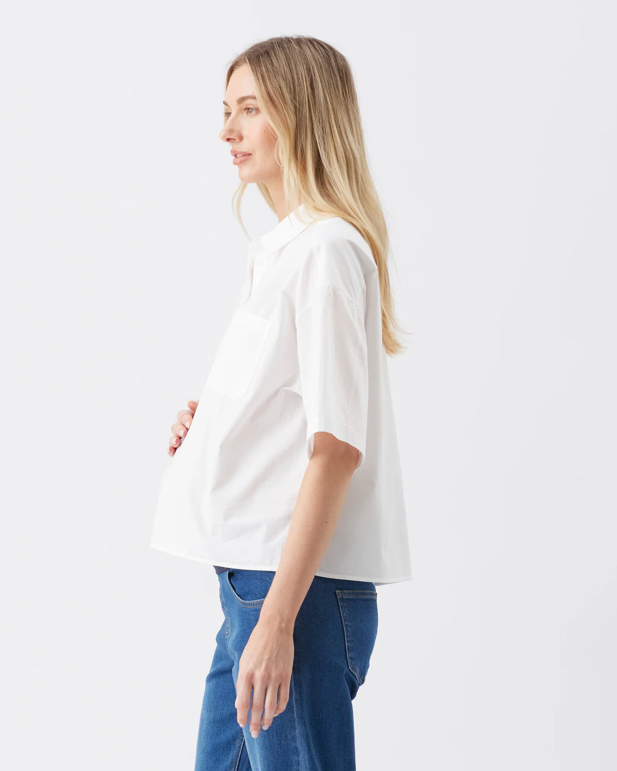 A pregnant woman shown in profile wearing a white short-sleeve maternity top and blue jeans against a plain white studio background.