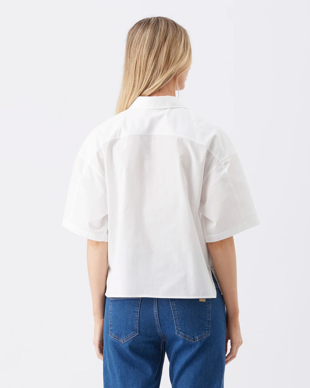 A woman viewed from behind wearing a white short-sleeve button-up shirt and blue jeans against a plain light gray studio background.