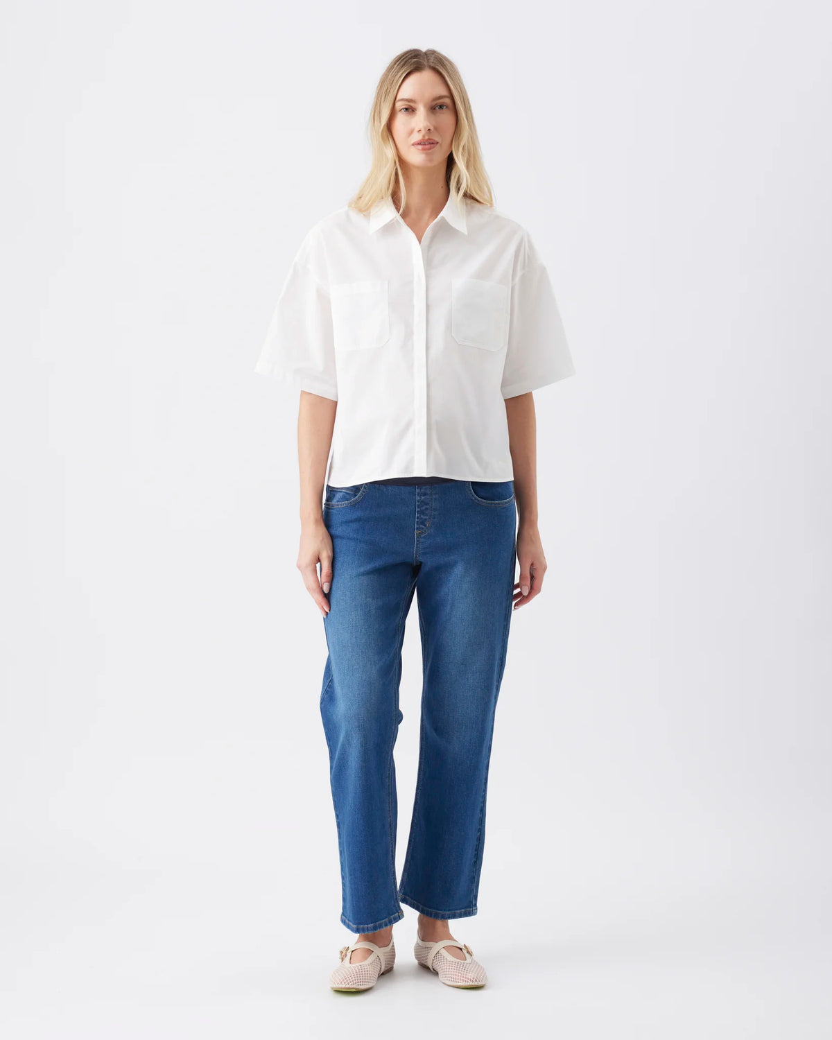 A woman stands against a plain light gray studio background wearing a white short-sleeve button-up shirt, blue jeans, and light beige shoes.